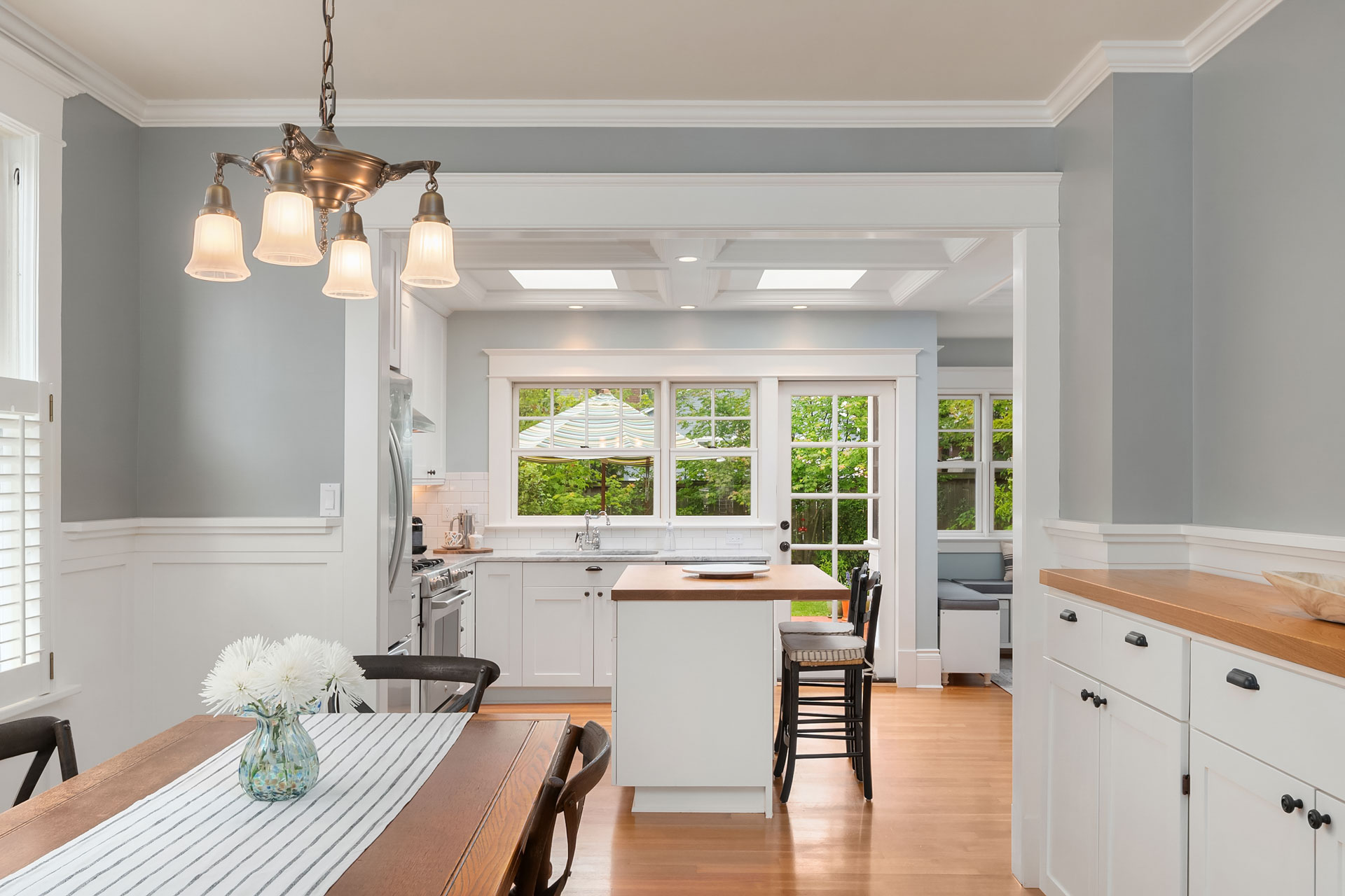 A bright, modern kitchen with white cabinets and a central island with black barstools. A dining table with a striped runner and a vase of flowers is in the foreground. The room is well-lit with ceiling lights and large windows offering a view of greenery outside.