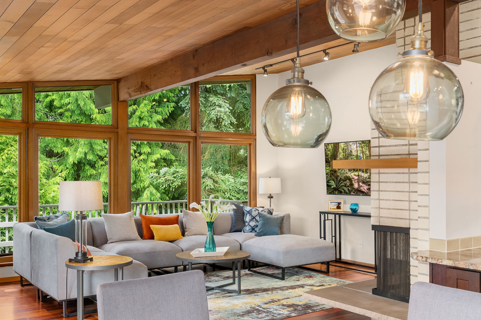 A contemporary living room with large windows showcasing a forest view. The room features a gray sectional sofa with colorful throw pillows, a modern wooden coffee table, and stylish hanging globe lights. A brick fireplace and modern decor enhance the cozy ambiance.