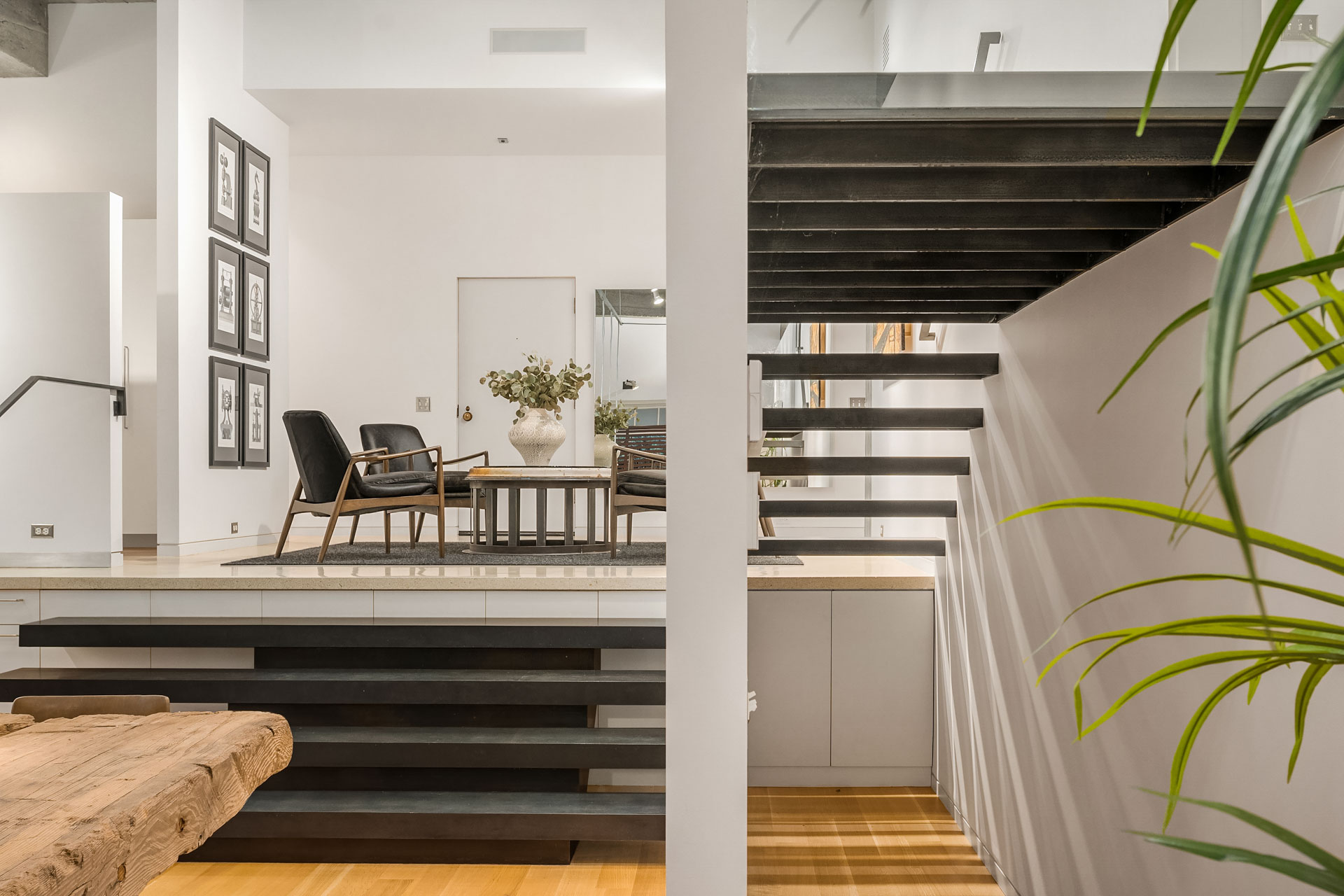 A modern interior featuring a metal floating staircase leading to a bright living area with two black armchairs, a wooden coffee table, and a framed photo collage. Wooden bench and green plant in the foreground, with minimalist décor throughout the space.