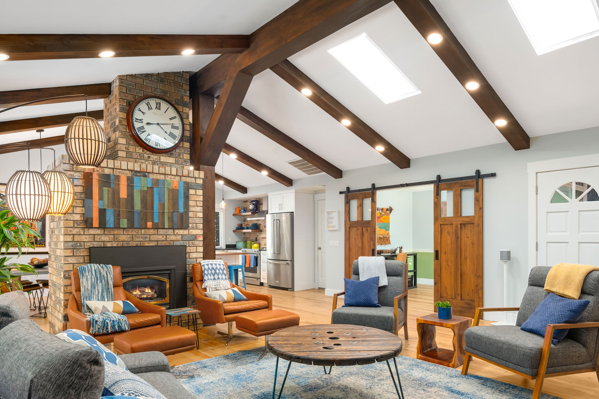 A cozy living room with vaulted ceilings featuring wooden beams and skylights. A brick fireplace with a clock above it is central, surrounded by comfortable seating. Sliding barn doors lead to another room. The space is decorated with modern and rustic elements.