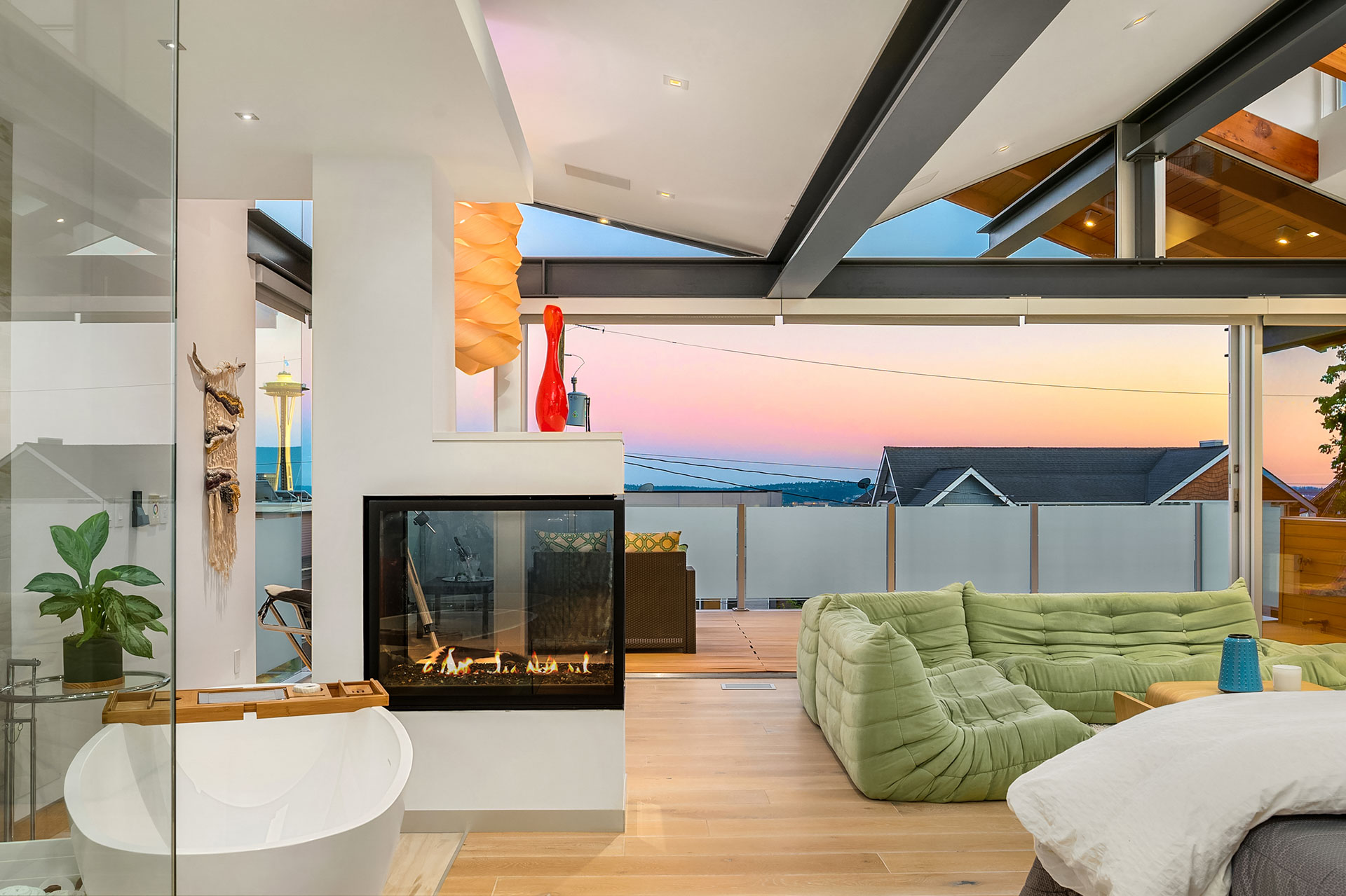 A modern, stylish open-plan living space with a cozy green sectional sofa by a fireplace. The room features large glass windows with a view of a colorful sunset over a balcony. A bathtub is situated in the corner, and sleek decor adorns the area.