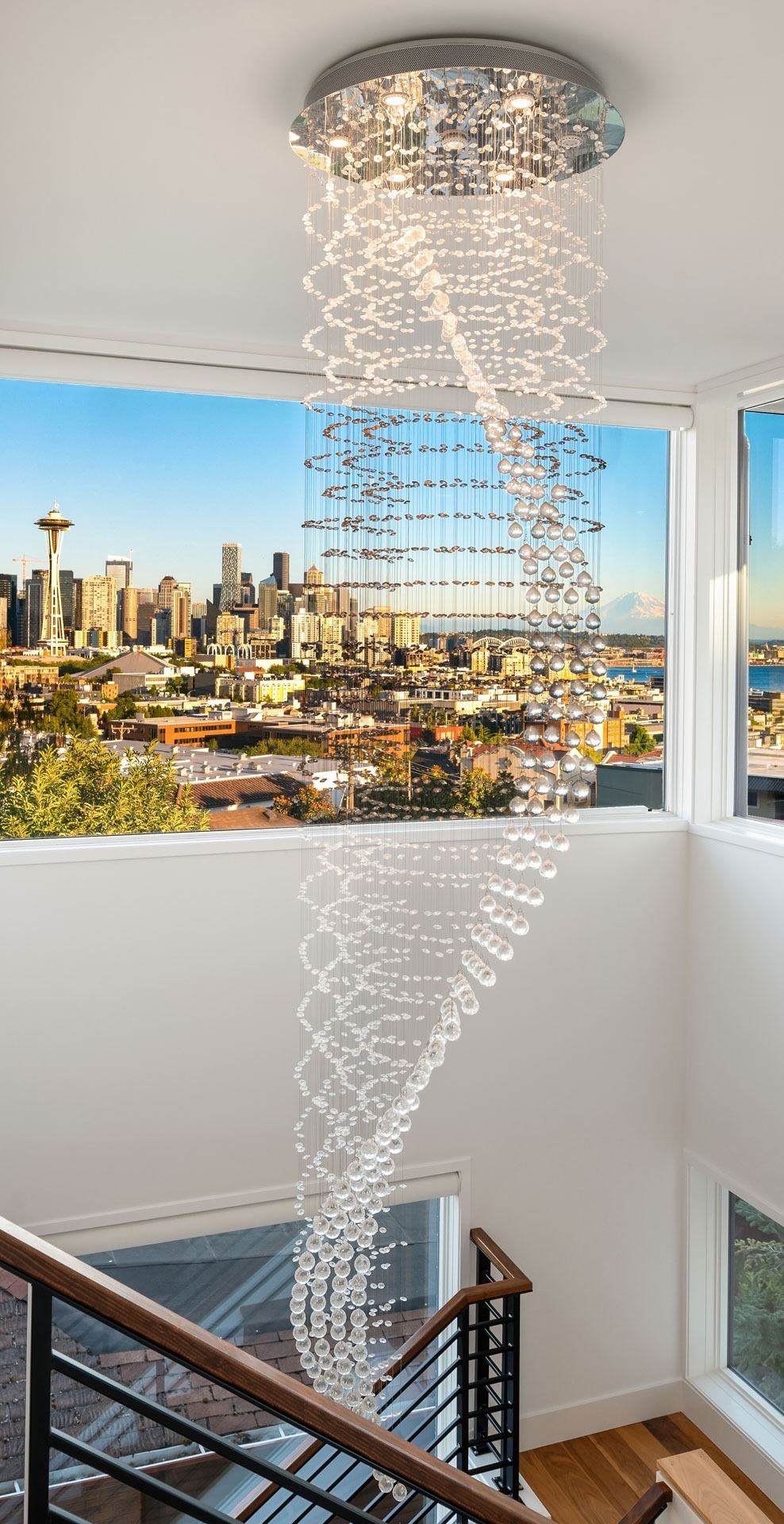 A modern chandelier with cascading glass pendants hangs in a stairwell with large windows. The windows offer a panoramic view of a city skyline, including the Space Needle, under a clear blue sky. A wooden handrail runs along the stairs.