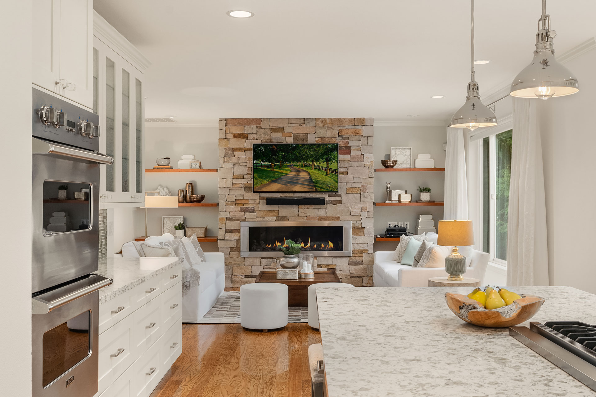 A modern, open-concept living room and kitchen. The living room has a stone fireplace, flat-screen TV, white sofas, and wooden shelves. The kitchen has stainless steel appliances, white cabinetry, a marble island, and pendant lights. A bowl of fruit sits on the island.