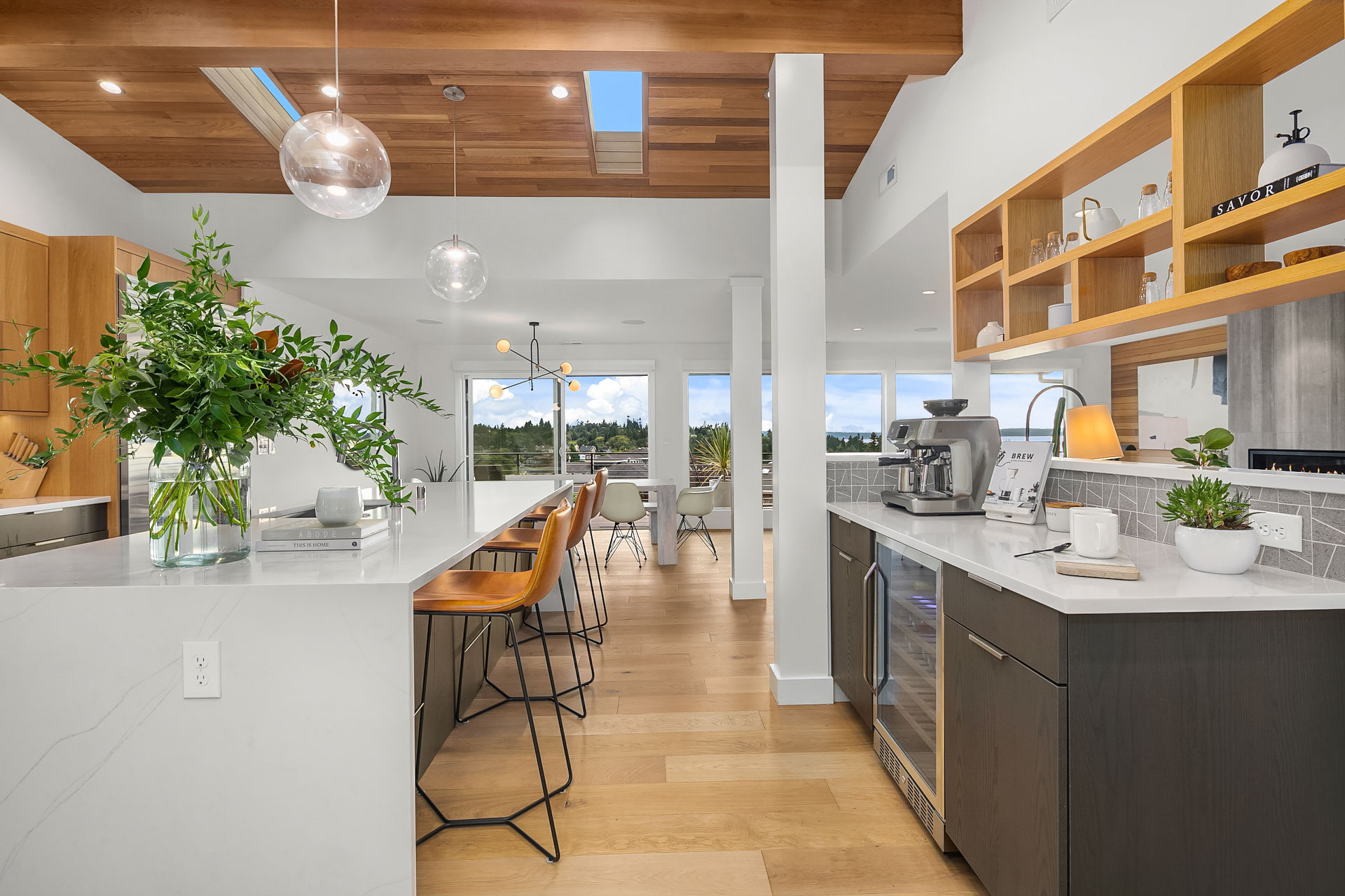 A modern kitchen featuring a white marble island with wooden bar stools, pendant lighting, and a high ceiling with wooden accents. The kitchen includes sleek appliances, open shelving with decor, and large windows offering a view of greenery.