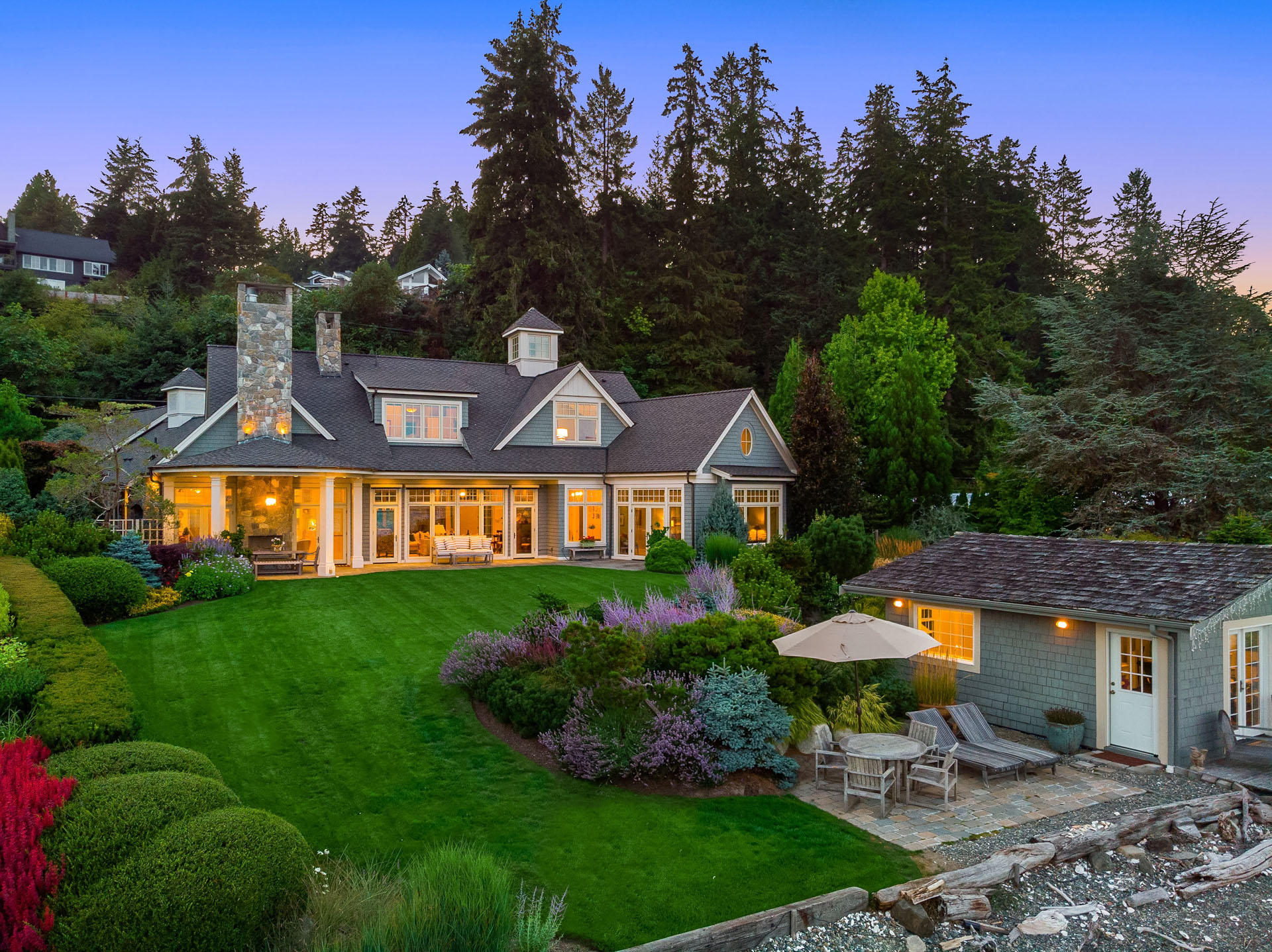 A large, two-story house with a stone chimney and multiple windows sits amid a lush green lawn and beautifully landscaped garden. The house features a wrap-around porch with outdoor seating, and a separate small structure with a patio area featuring a table and an umbrella. Dense trees provide a scenic backdrop.