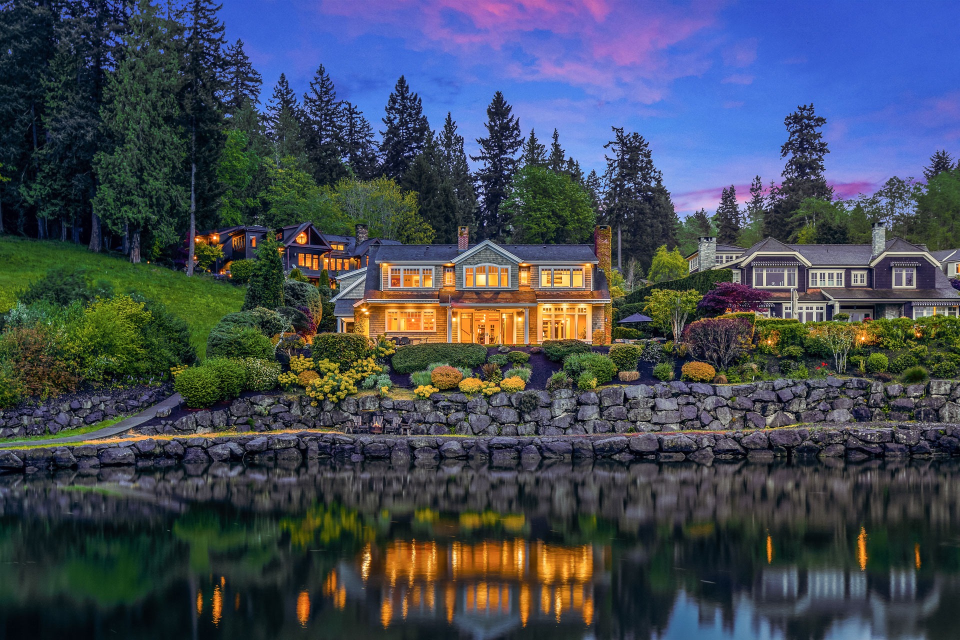 A beautiful, large house with many glowing windows at dusk, situated by a lake with its reflection visible in the water. The house is surrounded by lush greenery and manicured landscaping, with another illuminated house nearby. A backdrop of tall trees completes the scenic view.