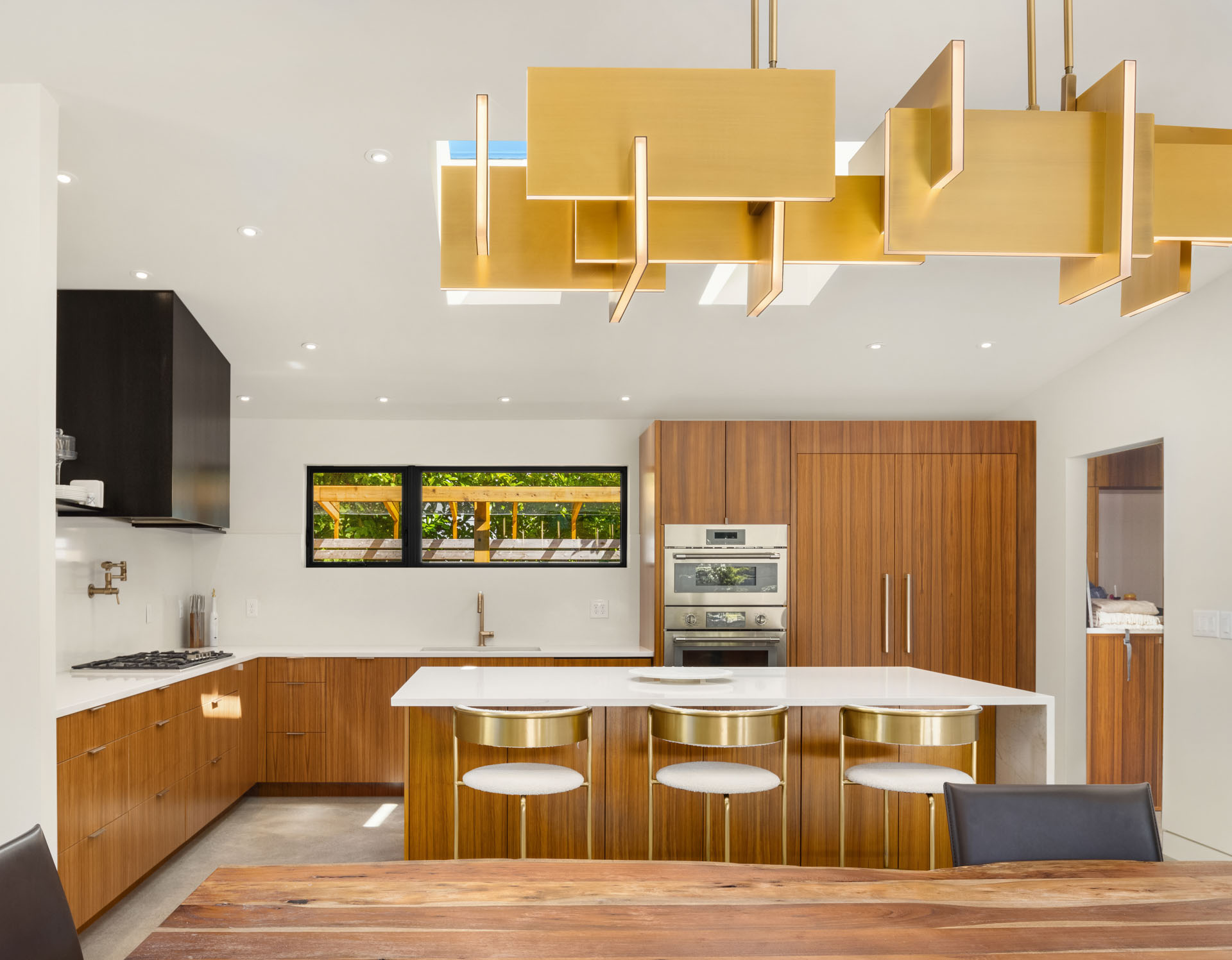 A modern kitchen features warm wooden cabinetry, a large white island with four contemporary bar stools, a black accented stove area, built-in stainless steel appliances, a geometric gold chandelier, and a window overlooking an outdoor space with greenery.