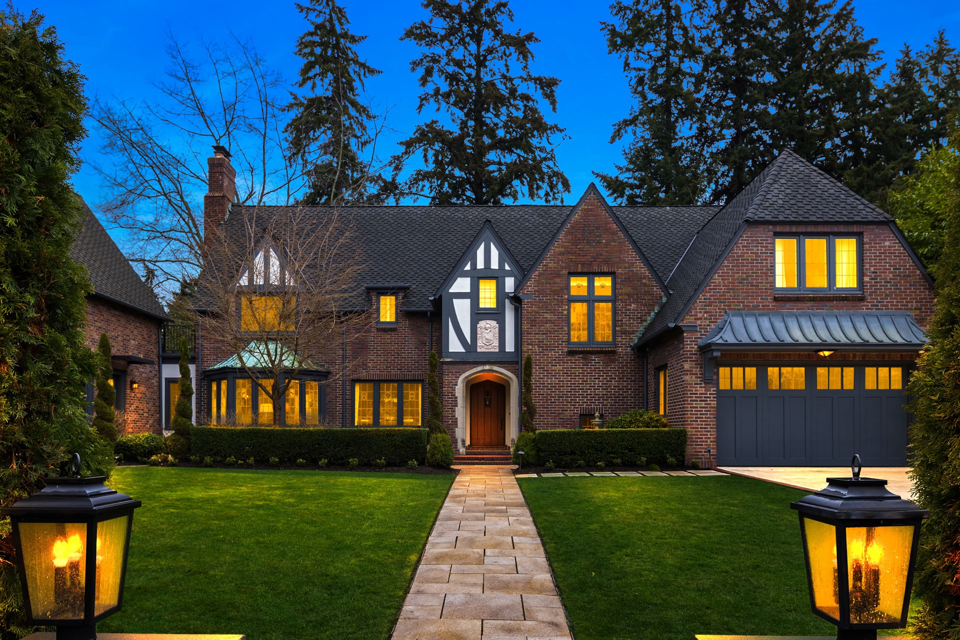 A large, two-story brick house with gabled roofs and black-and-white timber detailing. The home has numerous lit windows and a welcoming wooden front door. The house is surrounded by a well-manicured lawn, flanked by two illuminated lanterns, and framed by tall trees.