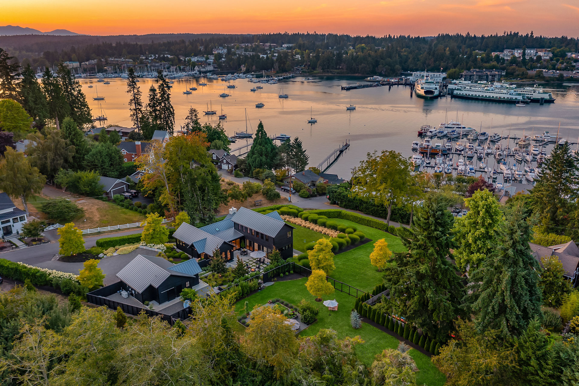 Aerial view of a scenic coastal town at sunset, with lush greenery and waterfront houses. A marina filled with boats is visible, along with a ferry docked nearby. The sky displays vibrant hues of orange and pink, reflecting on the calm water.