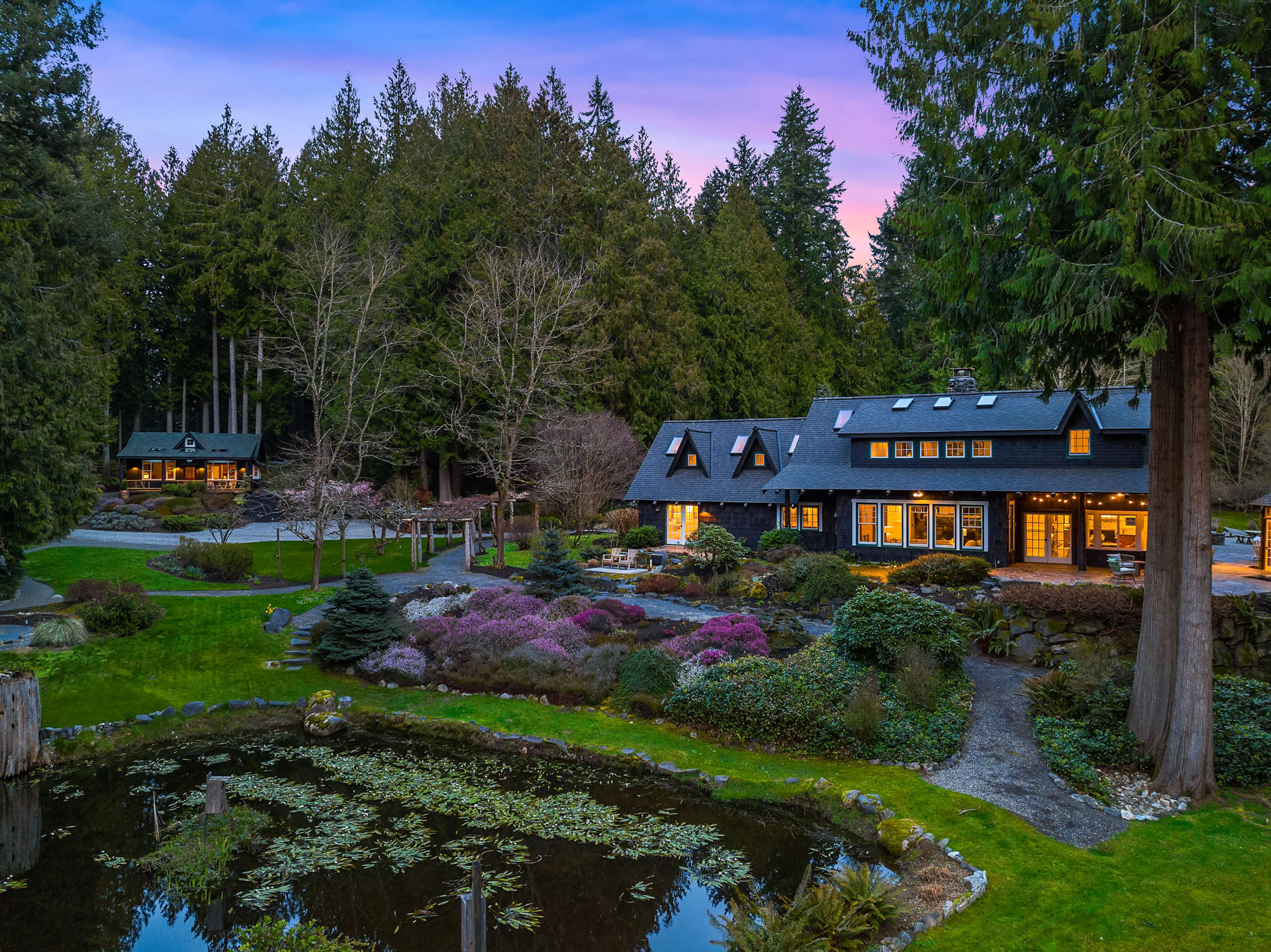 A picturesque property with a large, charming house surrounded by lush greenery and blooming gardens. There's a tranquil pond in the foreground with lily pads, and another smaller structure nestled among the tall trees in the background, all under a twilight sky.