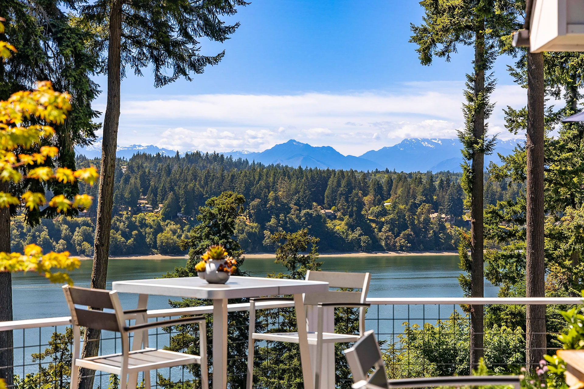 A scenic outdoor spot features a modern white table with chairs overlooking a serene lake, surrounded by tall trees. Mountains rise in the background under a clear blue sky, and small potted plants adorn the table, adding a touch of green.