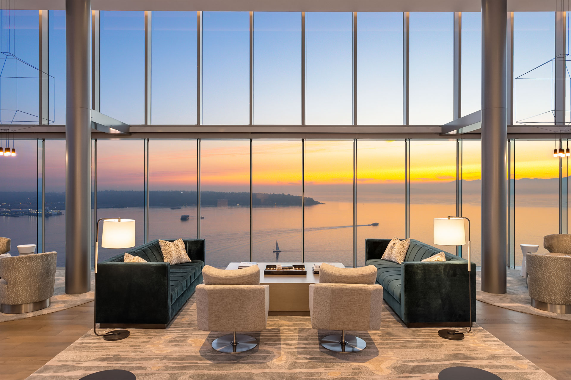 A luxurious, modern living room with floor-to-ceiling windows offers a panoramic view of a tranquil ocean at sunset. The room has two dark green sofas, two beige armchairs, and a stylish coffee table. Warm indoor lighting and neutral decor create a cozy atmosphere.