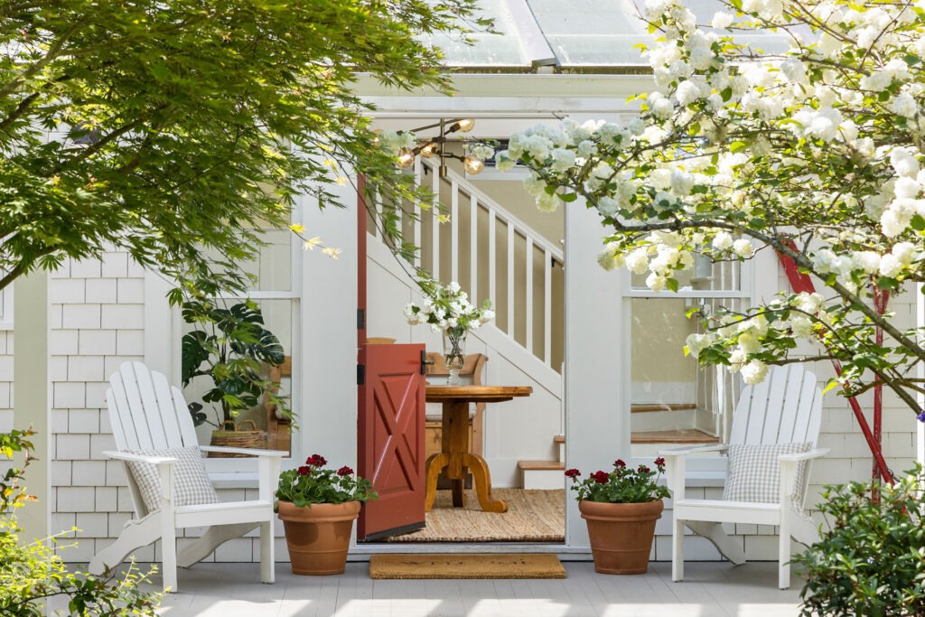 A charming outdoor seating area features two white Adirondack chairs with cushions, flanking a pair of terracotta planters filled with red flowers. An open red door leads to a bright interior with a wooden table and staircase, framed by blooming trees and greenery.