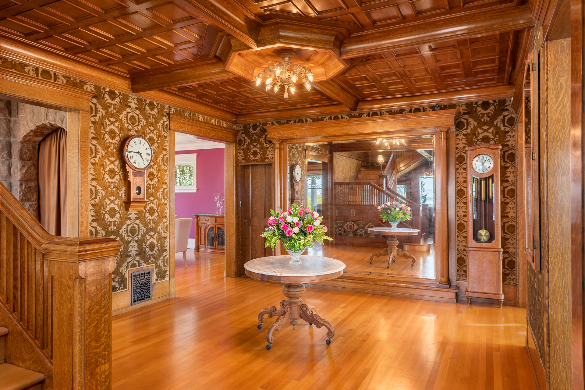 A grand hallway with ornate wallpaper features a wooden coffered ceiling and gleaming hardwood floors. A round marble table with a floral arrangement sits at the center. There is a large mirror on the back wall and an antique grandfather clock to the right.