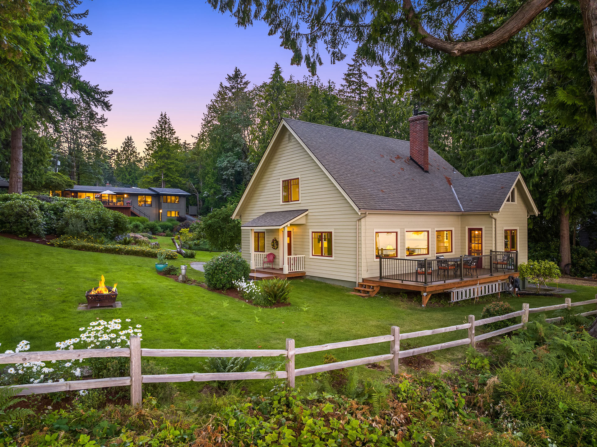 A charming yellow house with a steep gabled roof sits in a lush, green yard surrounded by trees. A wooden deck on the side has outdoor seating. In the background, another house is visible. A white picket fence and a small fire pit are in the foreground.