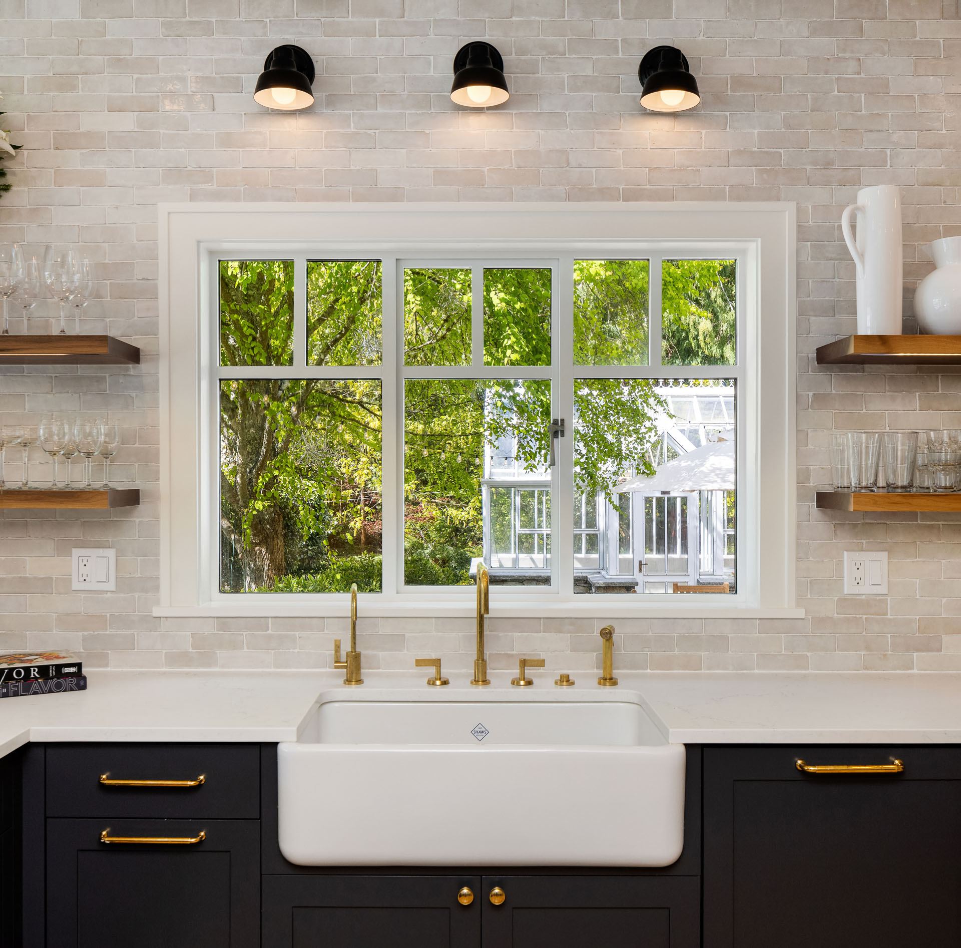 A kitchen features a large white farmhouse sink with brass fixtures below a wide window showing a green outdoor scene. Dark cabinets with brass handles, wooden shelves holding glassware and decor items, and three wall-mounted lights complete the stylish look.