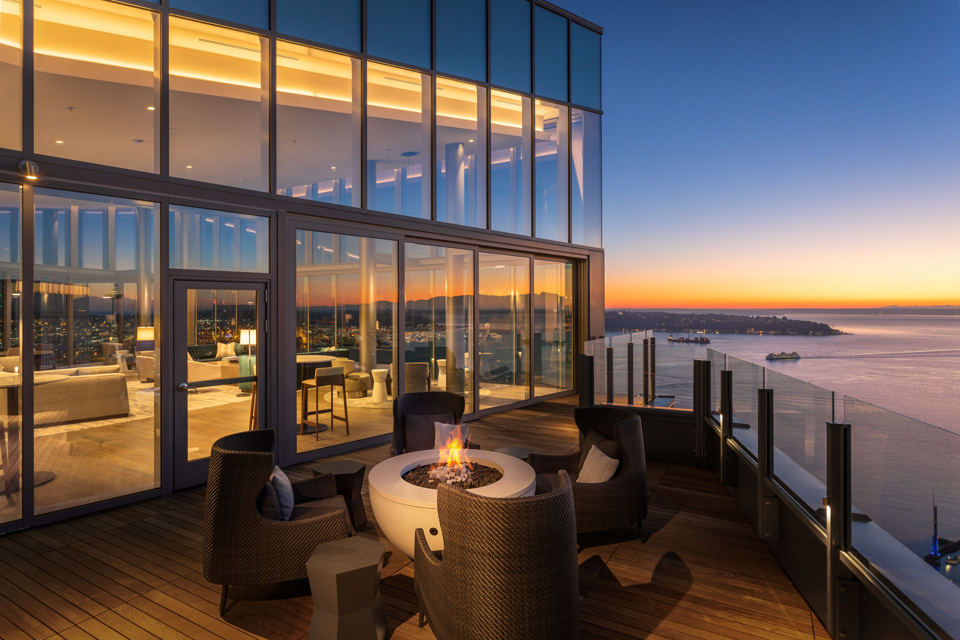 A modern rooftop lounge at sunset, featuring a cozy seating area around a fire pit. The lounge is enclosed by glass walls offering panoramic views of a city and waterfront below, with the sky transitioning from orange to deep blue as the sun sets.