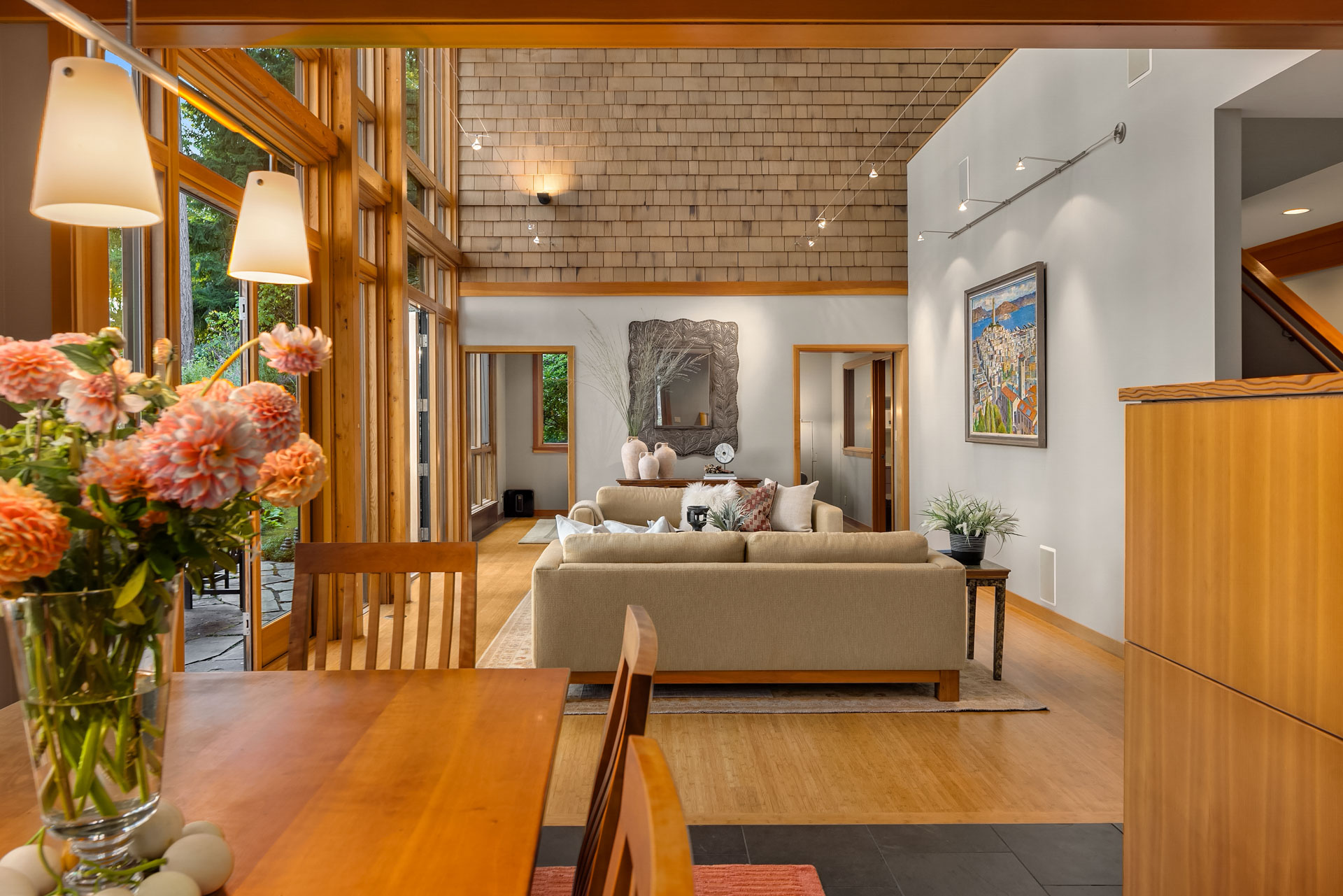 A modern living area with high ceilings and large windows, featuring a beige sofa, a wooden dining table with pink flowers, and stylish art pieces on the walls. The room is bright and has a mix of natural wood and soft neutral tones.