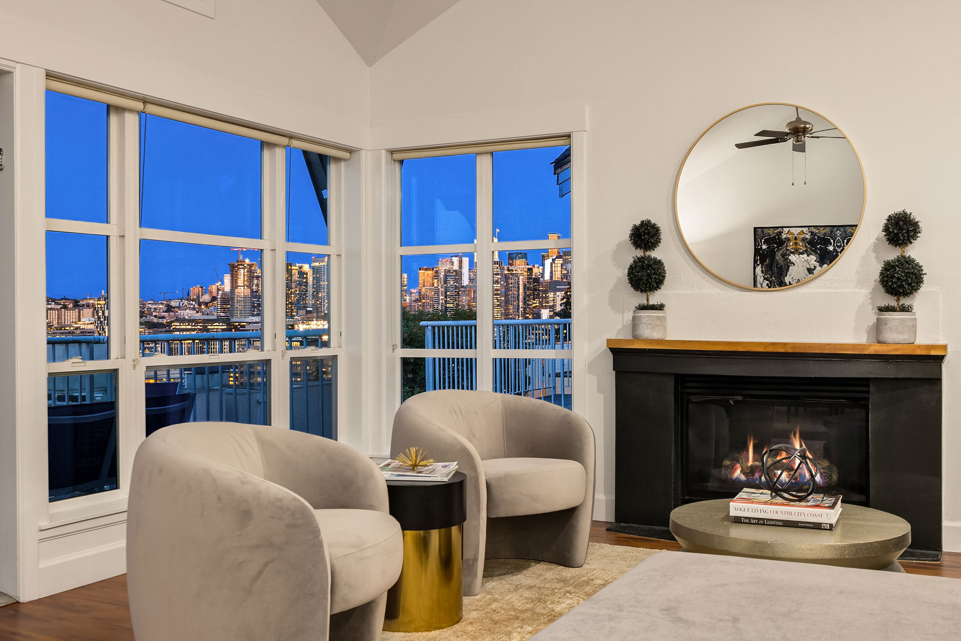 A modern living room features a cozy fireplace, two beige armchairs, a small round table with a gold base, and a mirror above the mantel. Large windows provide a view of a cityscape illuminated against a twilight sky. Decor includes potted plants and a wall art piece.