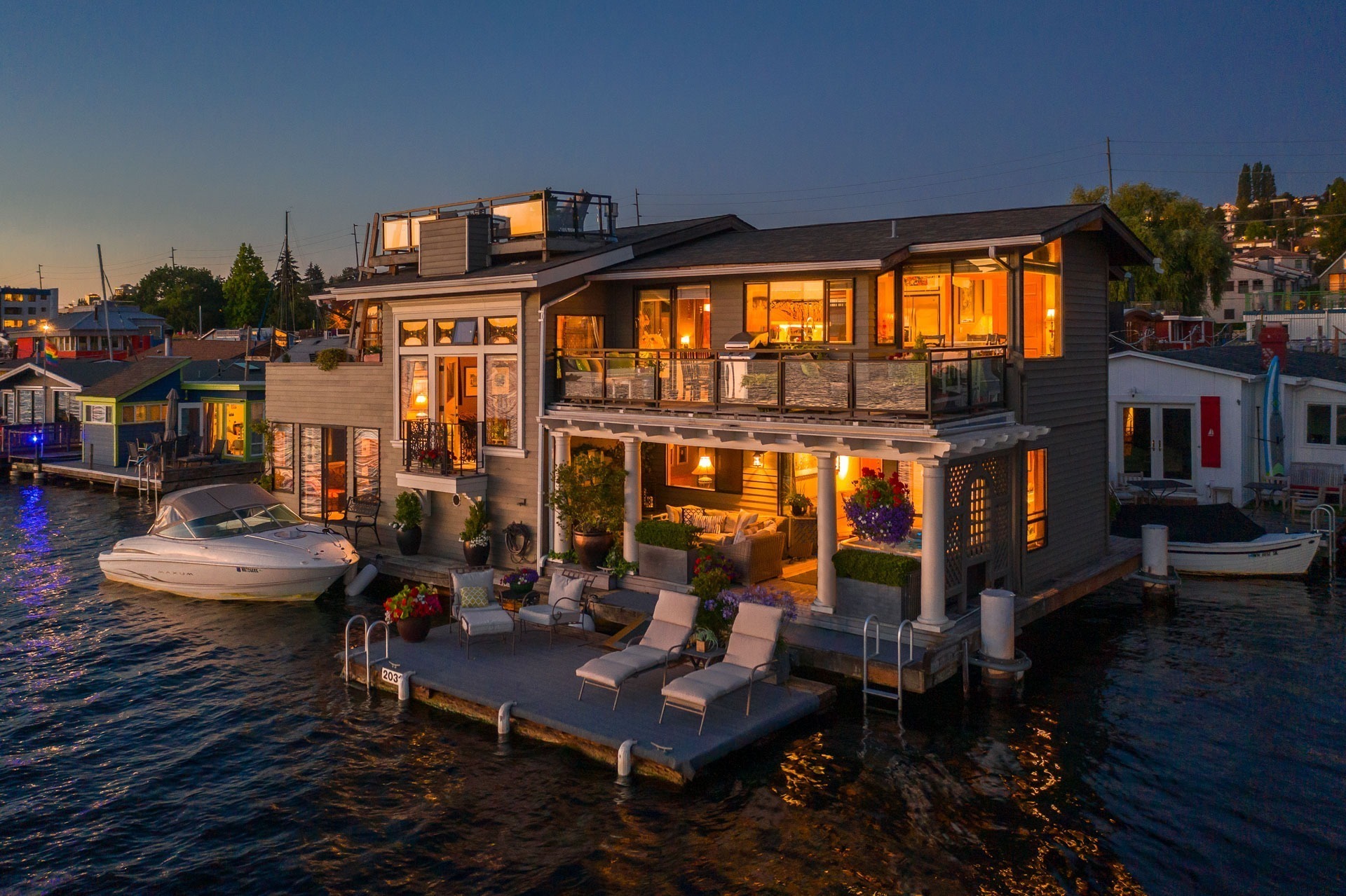 A picturesque evening view of a modern floating house on a calm body of water. The house is well-lit, exuding warmth, with large windows and multiple balconies. Surrounding the house, there are boats docked and adjacent homes similarly lit, creating a serene ambiance.