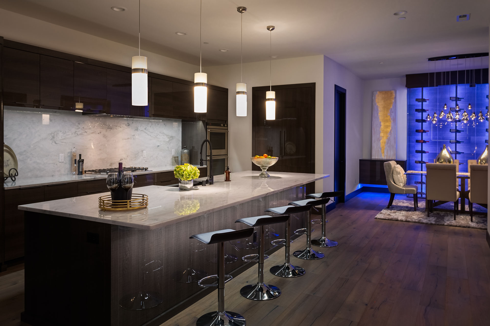 A modern kitchen and dining area with dark cabinetry, a large marble island with bar stools, and pendant lights. The dining area features contemporary furniture and blue accent lighting. A unique light fixture is above the dining table, and a wall art piece is illuminated.