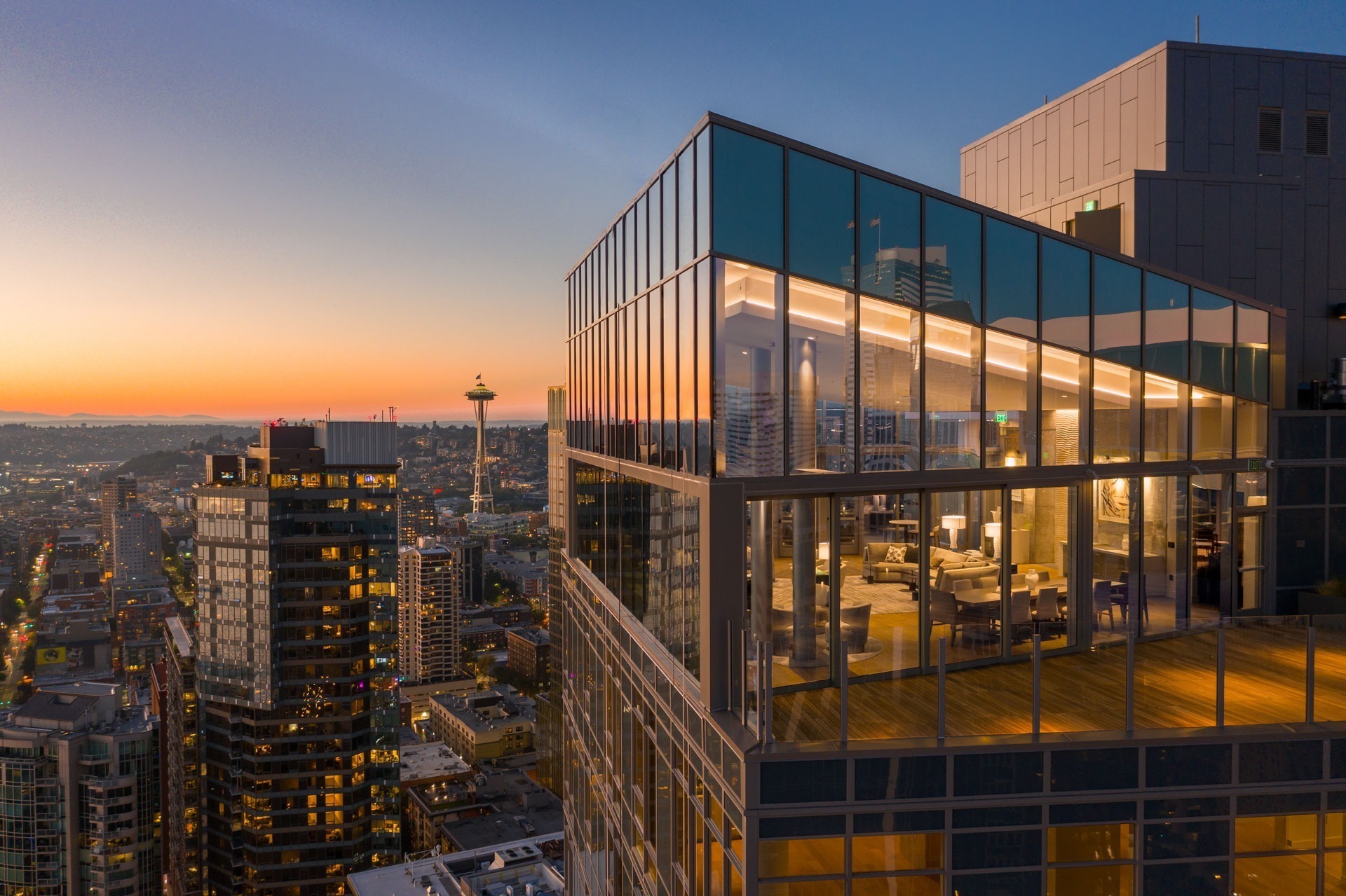 A luxury, modern glass apartment illuminated from within is perched atop a high-rise building at sunset. City buildings and the iconic Space Needle are visible in the background, under a colorful, evening sky.