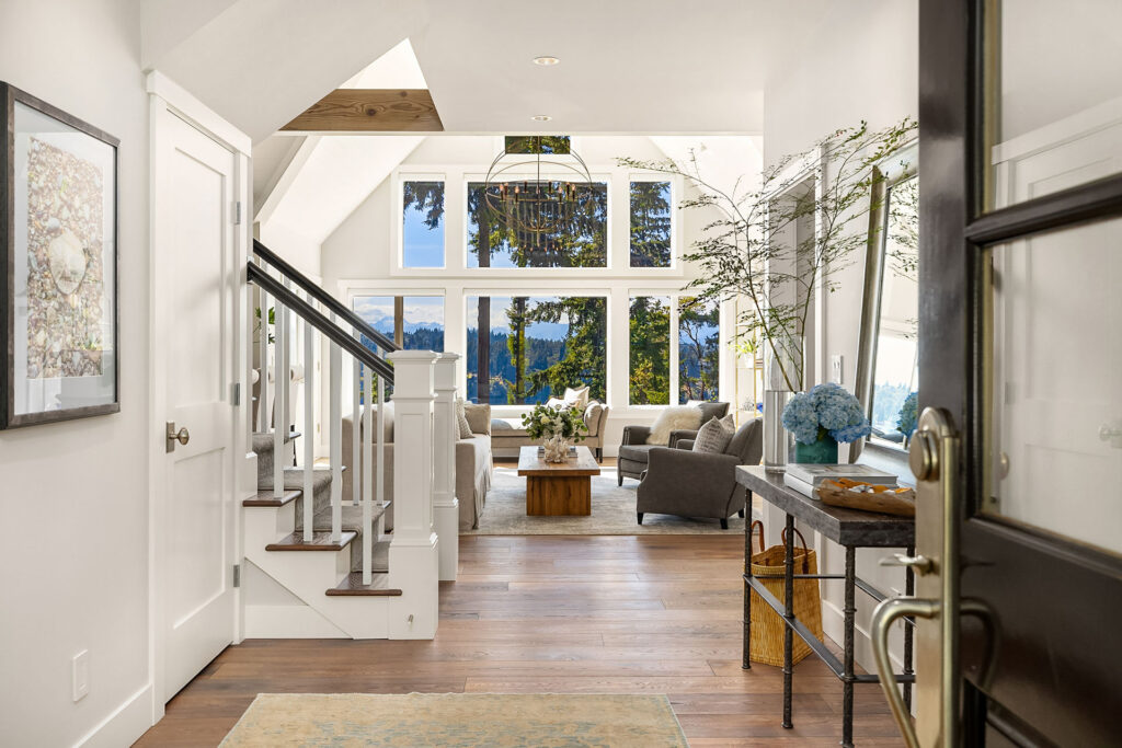 A welcoming foyer leads into a bright, spacious living room with tall windows offering a scenic view of trees and a lake. The space is tastefully decorated with modern furniture, a chandelier, and potted plants. A staircase is on the left, and a console table is on the right.