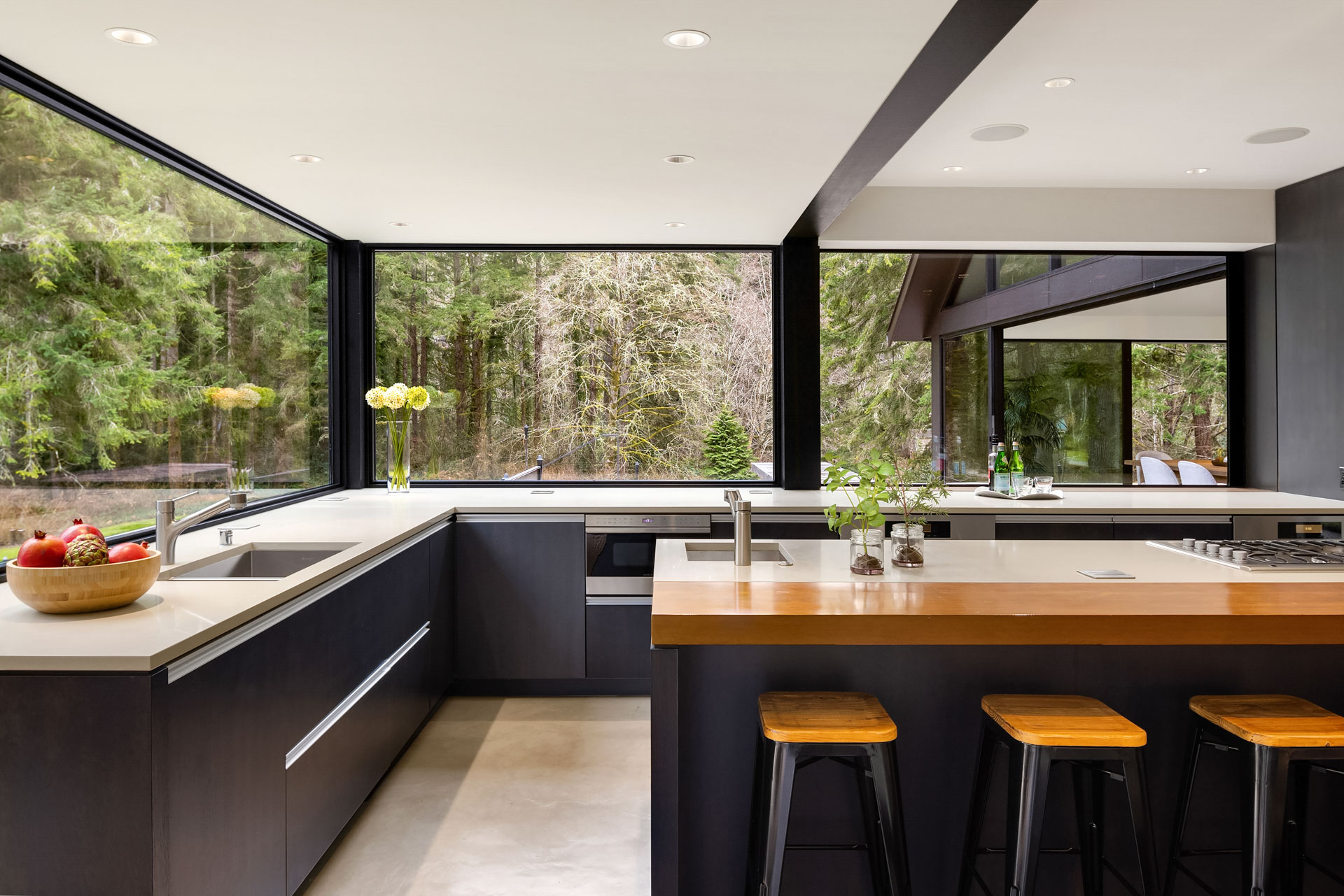 Modern kitchen with large windows providing a view of a lush forest. The kitchen features a sleek black and white design with wooden accents. It has an island with wooden stools, a bowl of fruit, and greenery placed around the room. Natural light brightens the space.