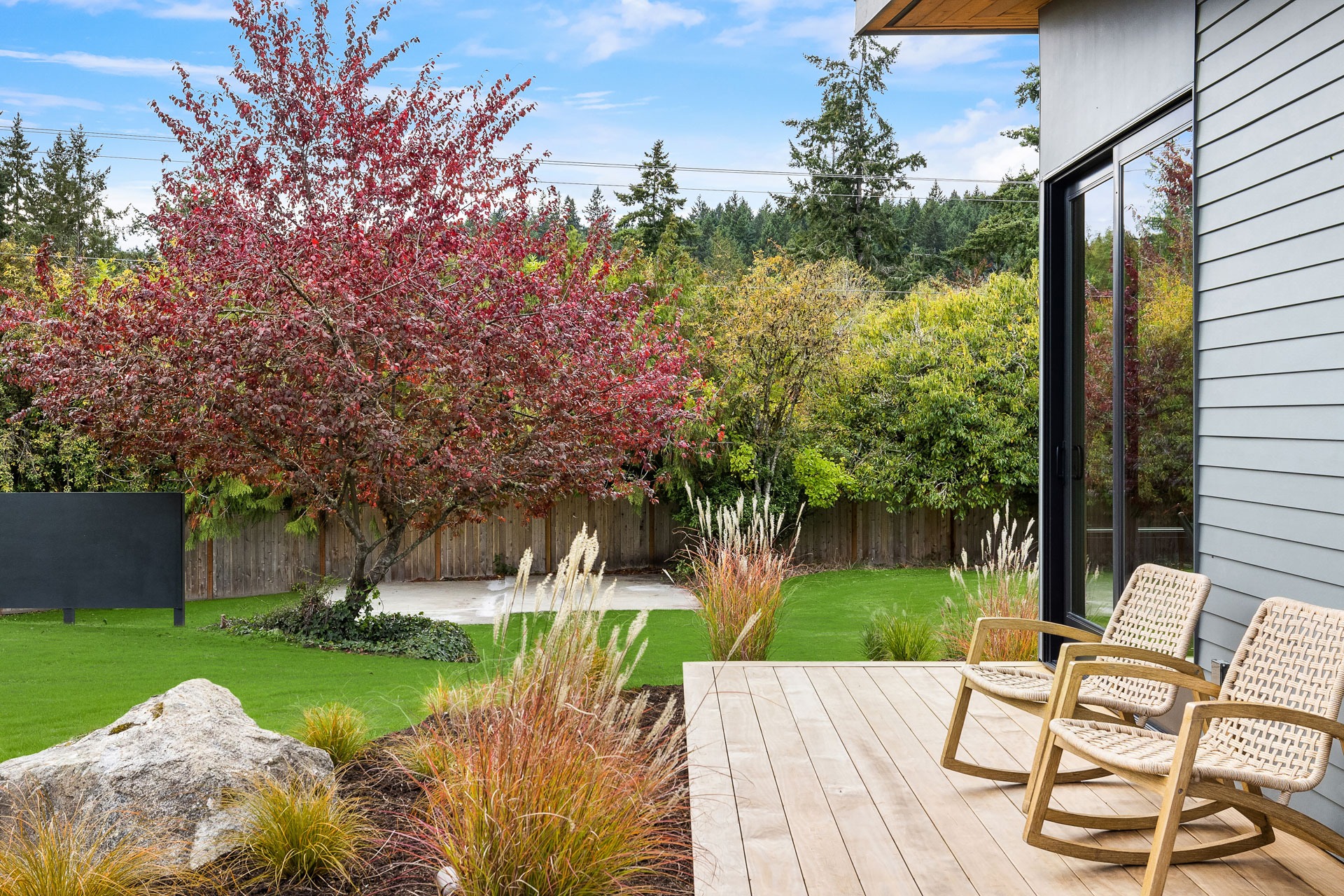 A modern backyard with a wooden deck featuring two wicker rocking chairs. The yard has green grass, landscaped plants, and a large tree with red leaves. A wooden fence borders the space, with additional greenery and a blue sky in the background.