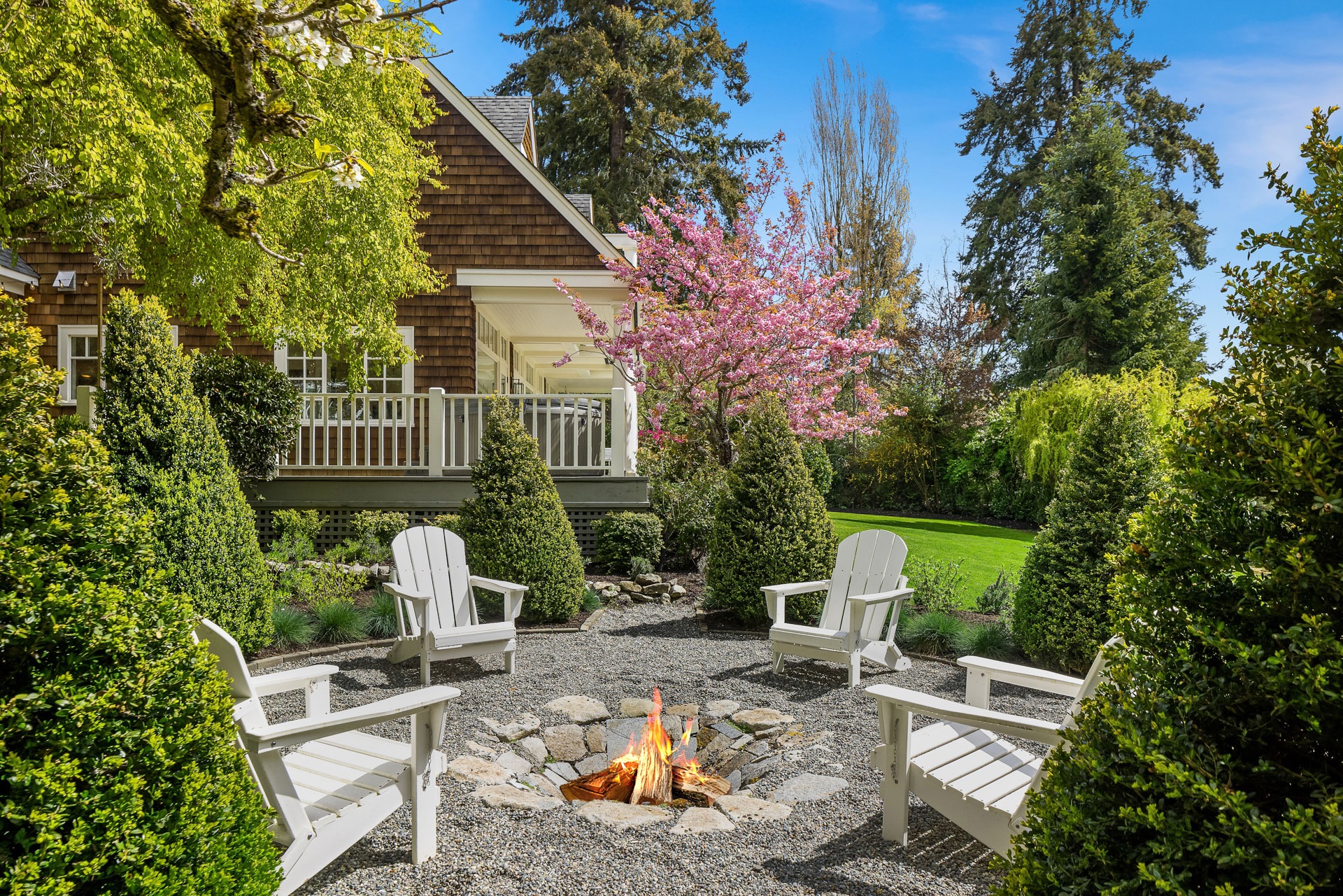 A cozy backyard patio features four Adirondack chairs arranged around a fire pit with a small stone base. The area is surrounded by lush greenery, hedges, blooming trees, and a well-manicured lawn. A charming house with a porch sits in the background under a clear blue sky.