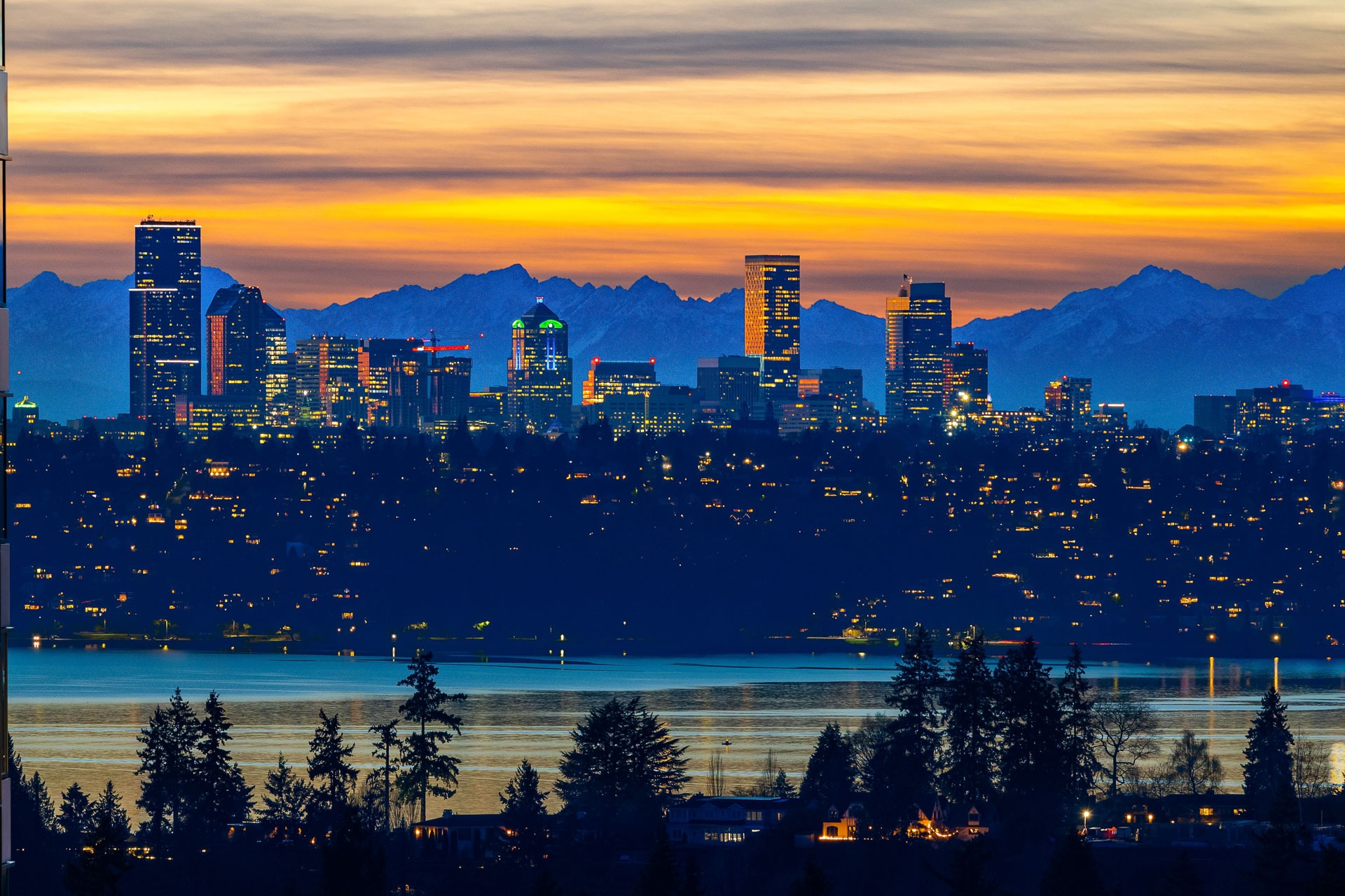 A stunning city skyline at dusk features tall buildings illuminated against a backdrop of a vibrant sunset, with hues of orange, yellow, and blue. Snow-capped mountains are visible in the distance, and a calm body of water reflects the city lights in the foreground.