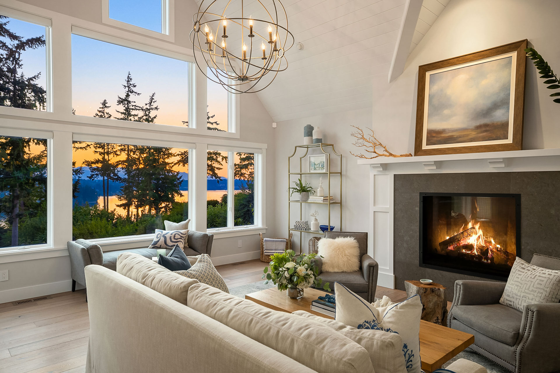 A modern living room with a fireplace, cozy seating, and a large window showcasing a scenic sunset over a water and forest landscape. The room has elegant decor, including a chandelier, a painting above the mantel, and a shelf with decorative items.