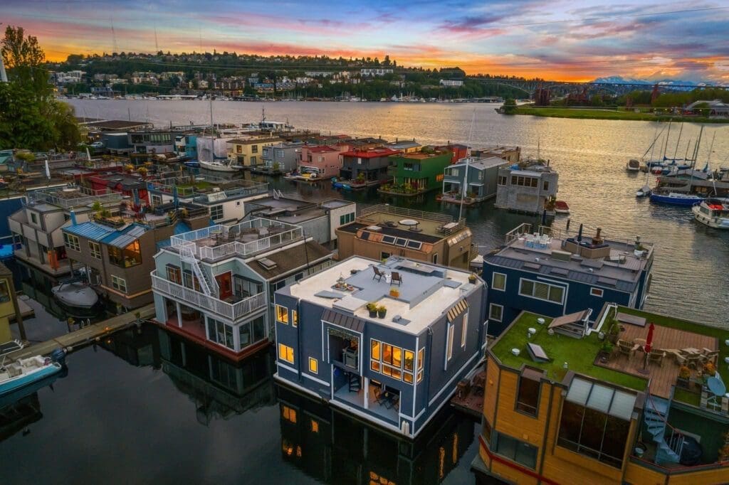 Real estate aerial photography view of colorful houseboats on a tranquil lake at sunset. The sky is painted with shades of orange, pink, and blue. Trees and buildings are seen in the distance, and several boats are docked along the shore. The houseboats have varying designs and rooftop decks.