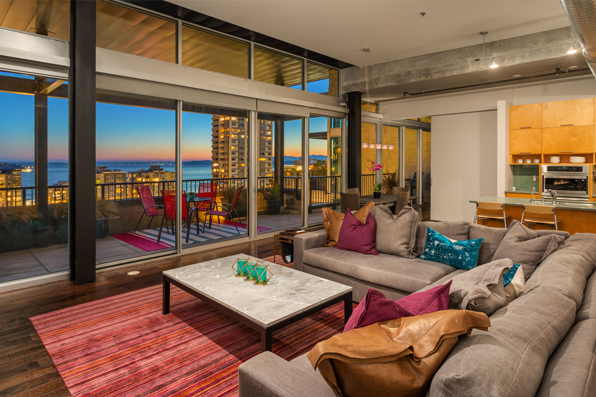 A modern apartment living room with floor-to-ceiling windows opens to a balcony offering a cityscape view at sunset. The room features a gray sectional sofa with colorful pillows, a marble-top coffee table on a striped rug, and a kitchen area with wooden cabinets.