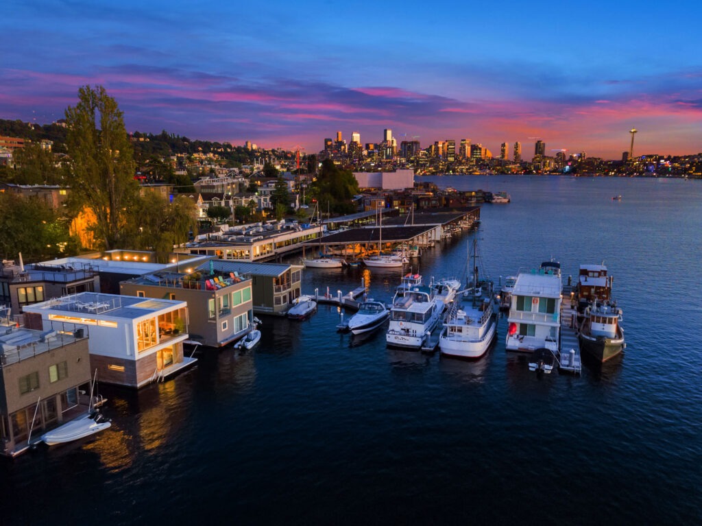 A vibrant cityscape at dusk with boats docked at the lakeside marina in the foreground. Modern houseboats and yachts line the shore. The illuminated skyline and a distinctive observation tower are visible against a colorful sunset sky.