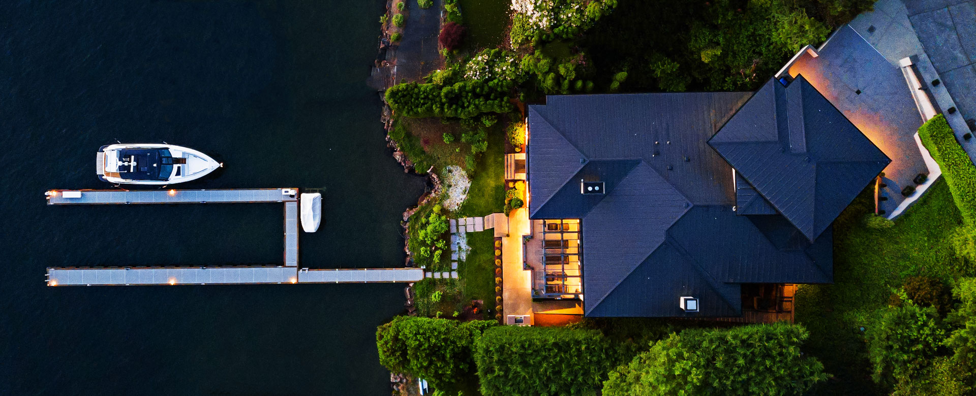 Aerial view of a luxurious lakeside property with a large modern house surrounded by greenery. The house features multiple patios and warm outdoor lighting. A lengthy dock extends out onto the water, where a white boat is moored.