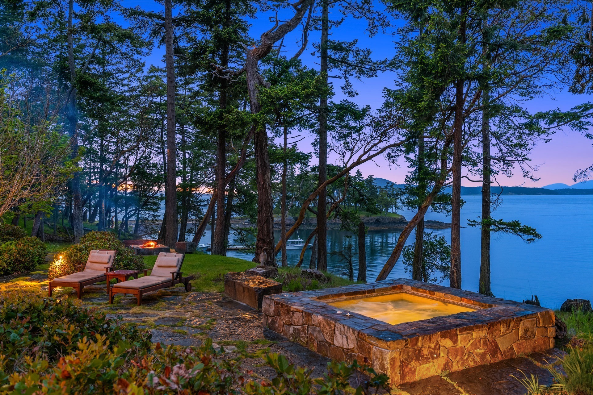 A serene outdoor setting features two lounge chairs on a stone patio near a hot tub surrounded by nature. Tall trees and lush greenery overlook a calm, expansive body of water, with a soft, colorful evening sky in the distance.