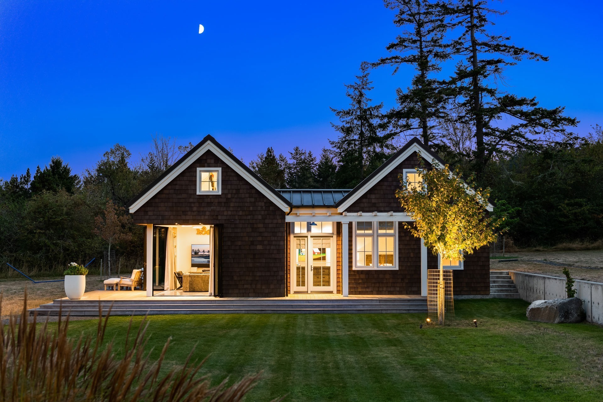 A charming, dark brown cottage with illuminated windows stands amidst a landscaped lawn at dusk. A small tree with a spotlight and an outdoor seating area enhance the cozy atmosphere. The clear sky overhead features a crescent moon, and tall trees frame the background.