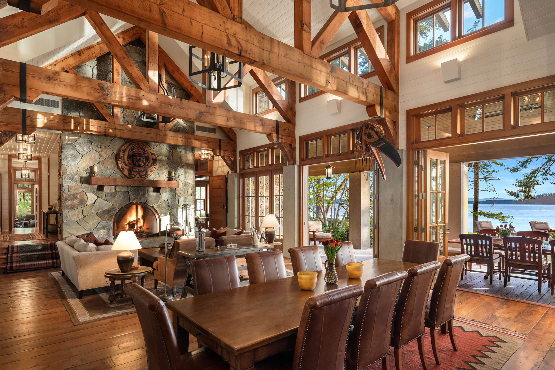 A spacious, rustic living and dining area with exposed wooden beams, stone fireplace, and large windows providing abundant natural light. The room features leather furniture, a wooden dining table, and views of a lake and trees outside.