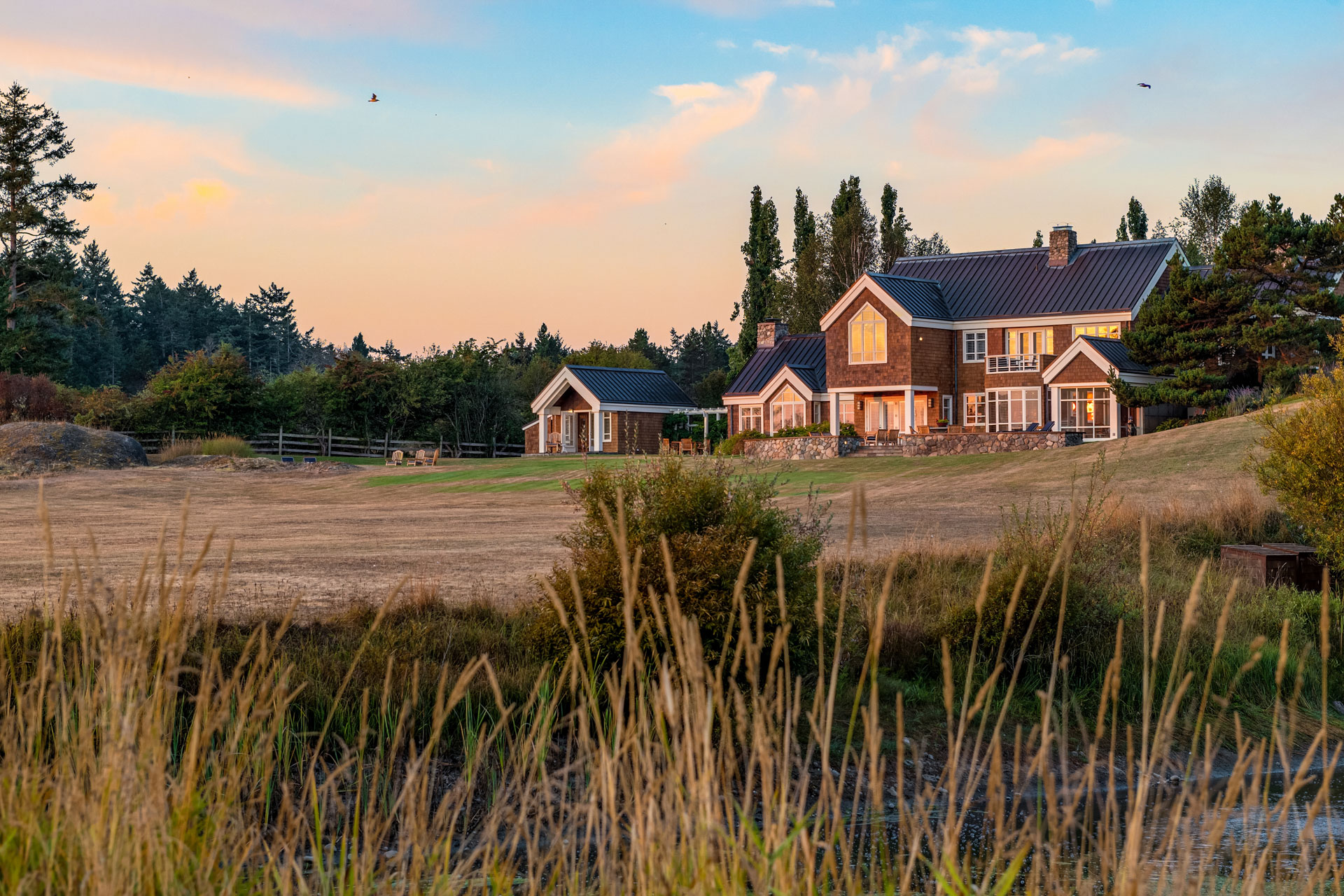 A scenic view of a large house amid lush greenery at sunset. Tall grasses in the foreground partially obscure the house. Trees surround the house, and a clear sky with a few flying birds is in the background. Warm lighting enhances the tranquil atmosphere.