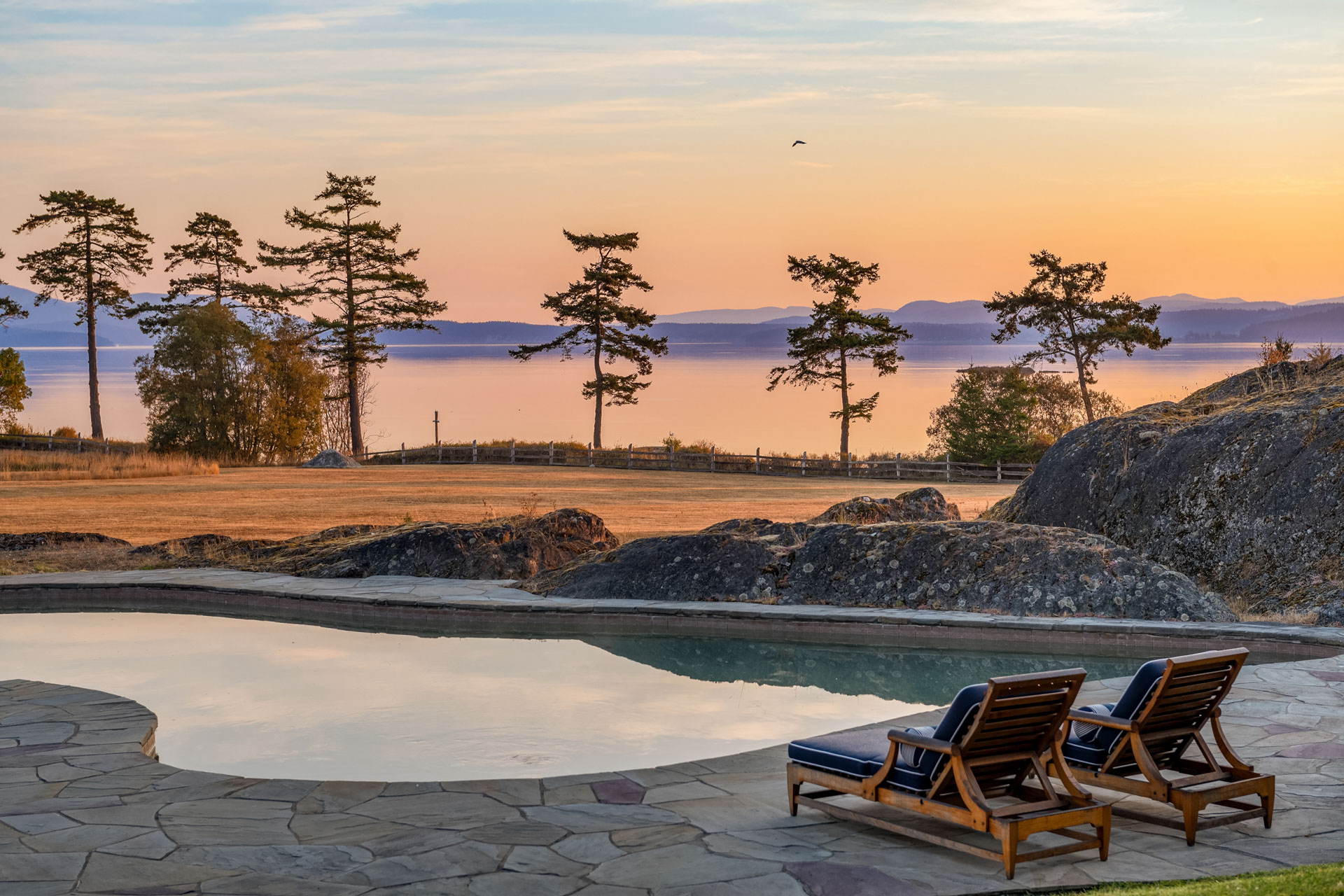 Two wooden lounge chairs with blue cushions sit by a pool, overlooking a scenic view of distant trees, a calm body of water, and a vibrant sunset. The pool is surrounded by a stone patio and natural rock formations, creating a serene and relaxing atmosphere.