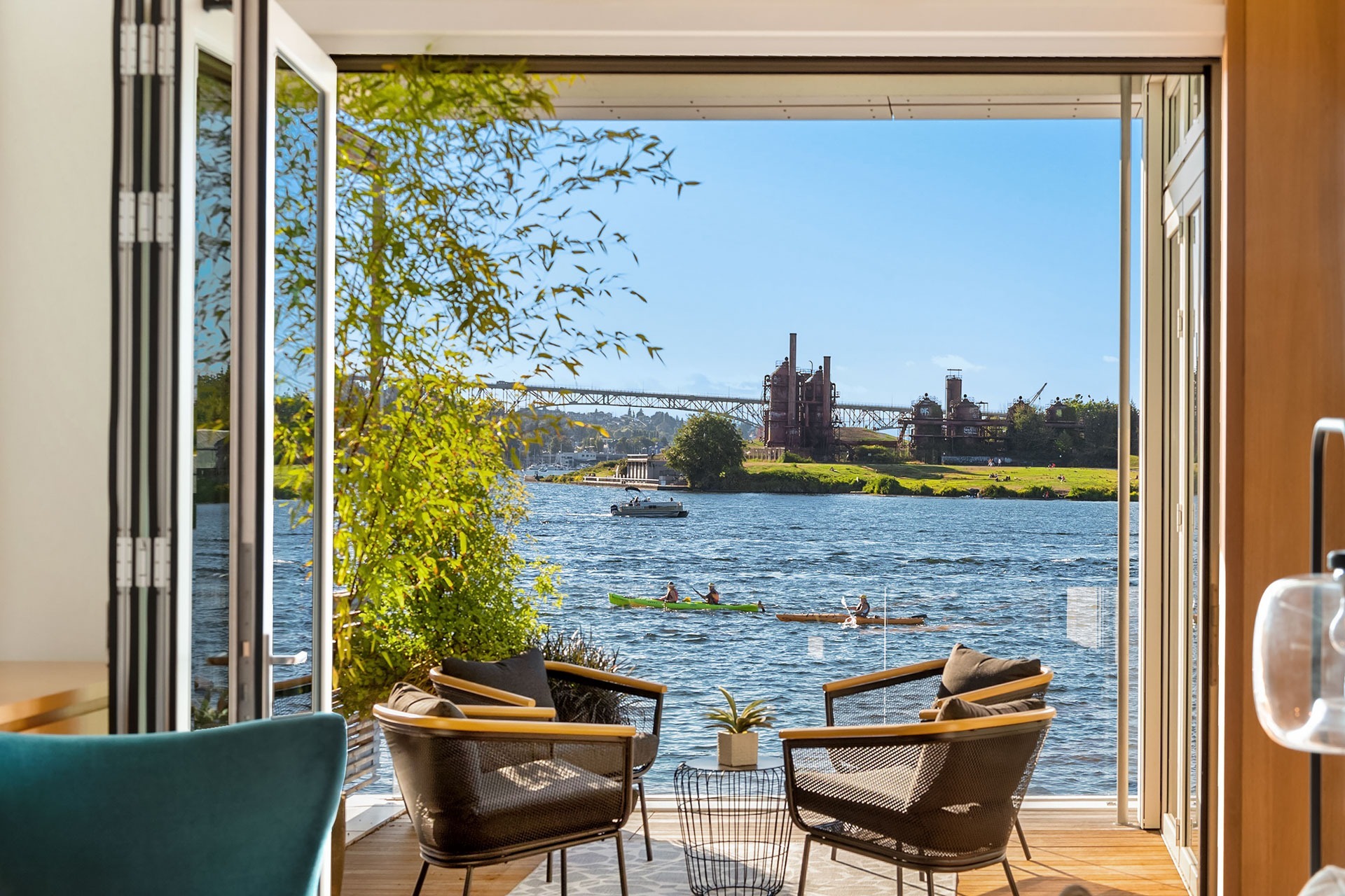 View from a modern home's interior featuring an open sliding door leading to a patio with two chairs and a table. Beyond, there's a body of water with rowers, a green landscape, an industrial structure in the distance, and a clear blue sky.