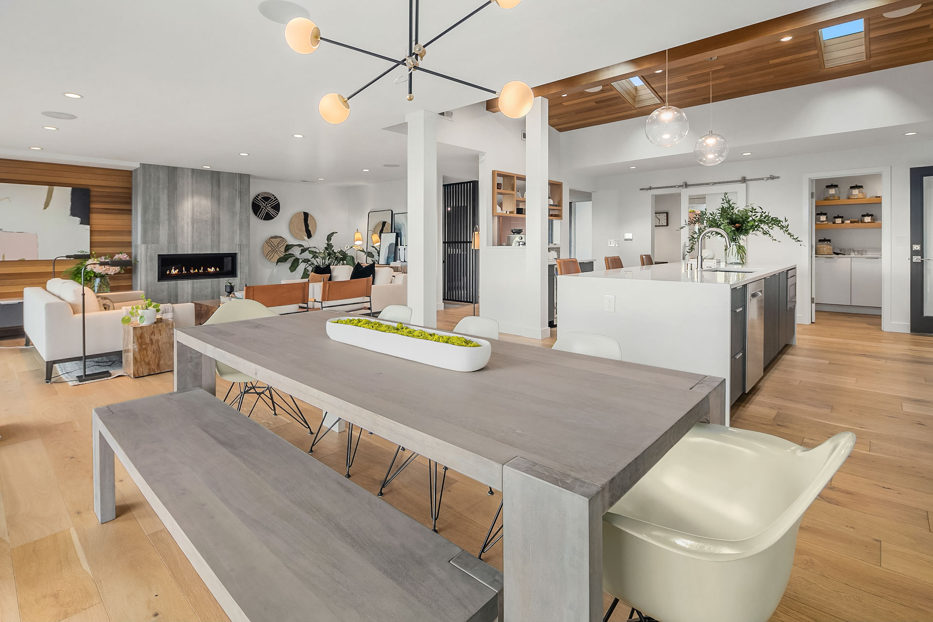 Luxurious modern open-concept living space with a dining area, living room, and kitchen. Features include a large wooden dining table with bench seating, a contemporary chandelier, sleek cabinetry, built-in fireplace, natural light, and minimalist decor.