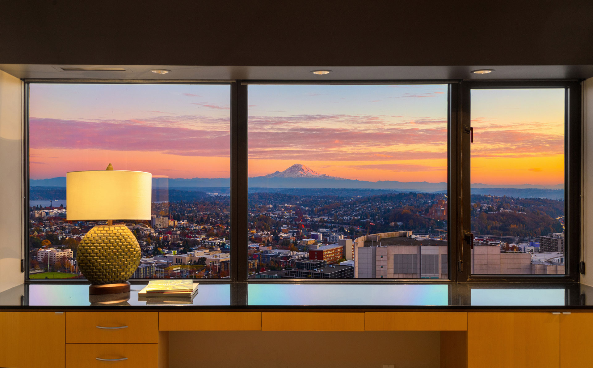 A view of a cityscape with buildings and streets at sunset, framed by a large window. The room features a countertop with a lamp and a few books. In the distance, a mountain is visible under a colorful sky with shades of pink, orange, and blue.