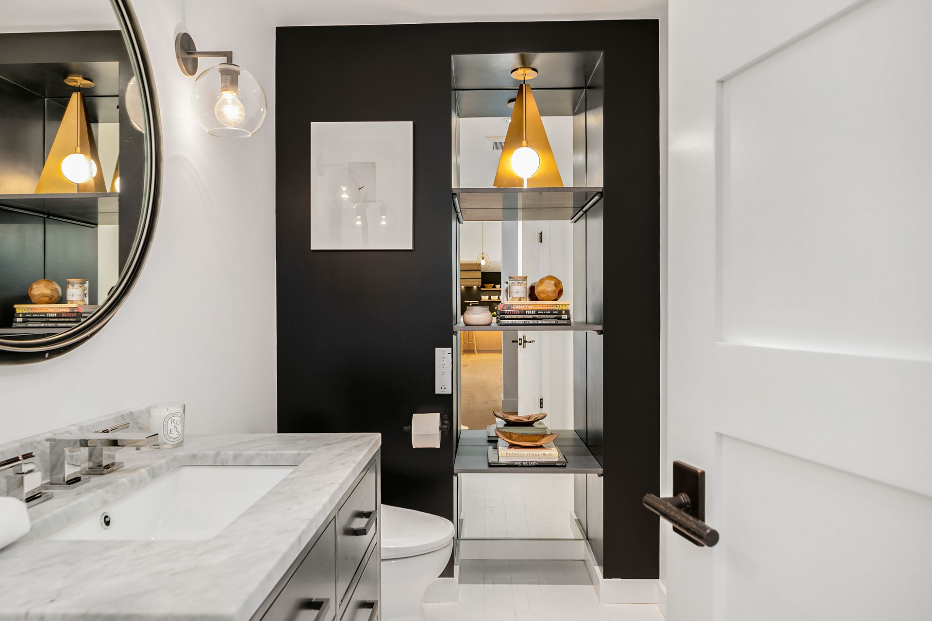 A modern bathroom features a white marble countertop with a mounted sink, a round mirror above, and a black accent wall with a sleek shelving unit. The shelves hold decorative items, and a golden pendant light hangs from the ceiling, illuminating the space.