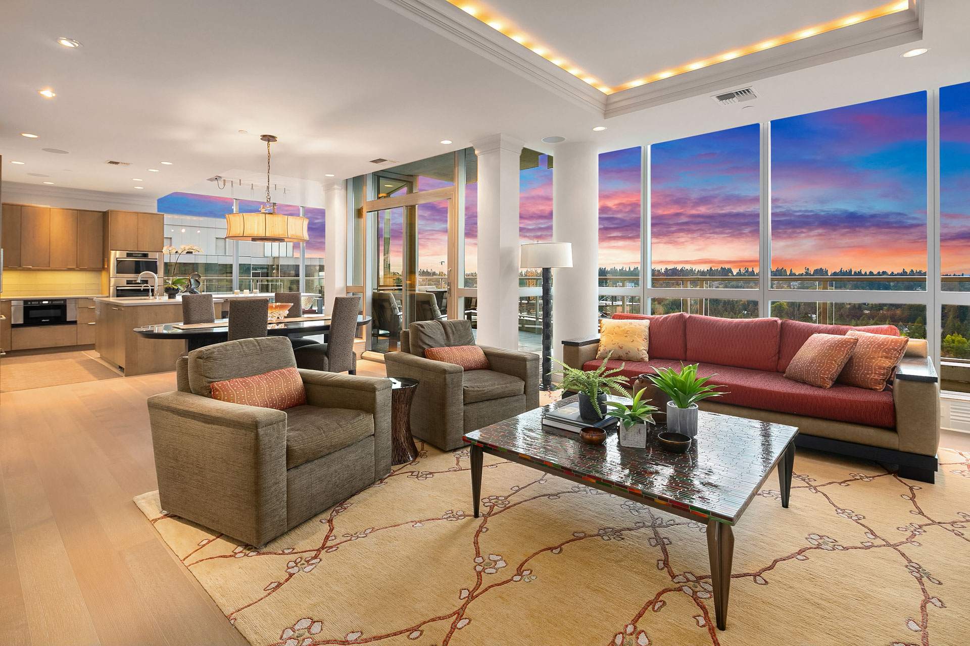 Luxurious open-concept living room and kitchen with modern furnishings. Large windows reveal a vibrant sunset over a forested landscape. The space features a red sofa, two armchairs, a glass coffee table with plants, and a chandelier above a dining area.