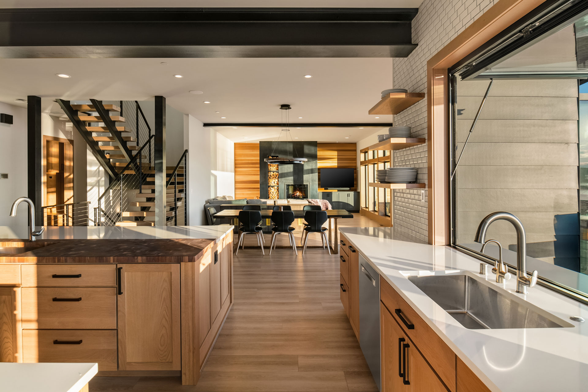 Modern kitchen with light wood cabinetry and white countertops. A large island with a sink sits in the foreground. Open shelving is visible on the right wall. In the background, there is a dining table, black chairs, and a living area with a staircase to the left.