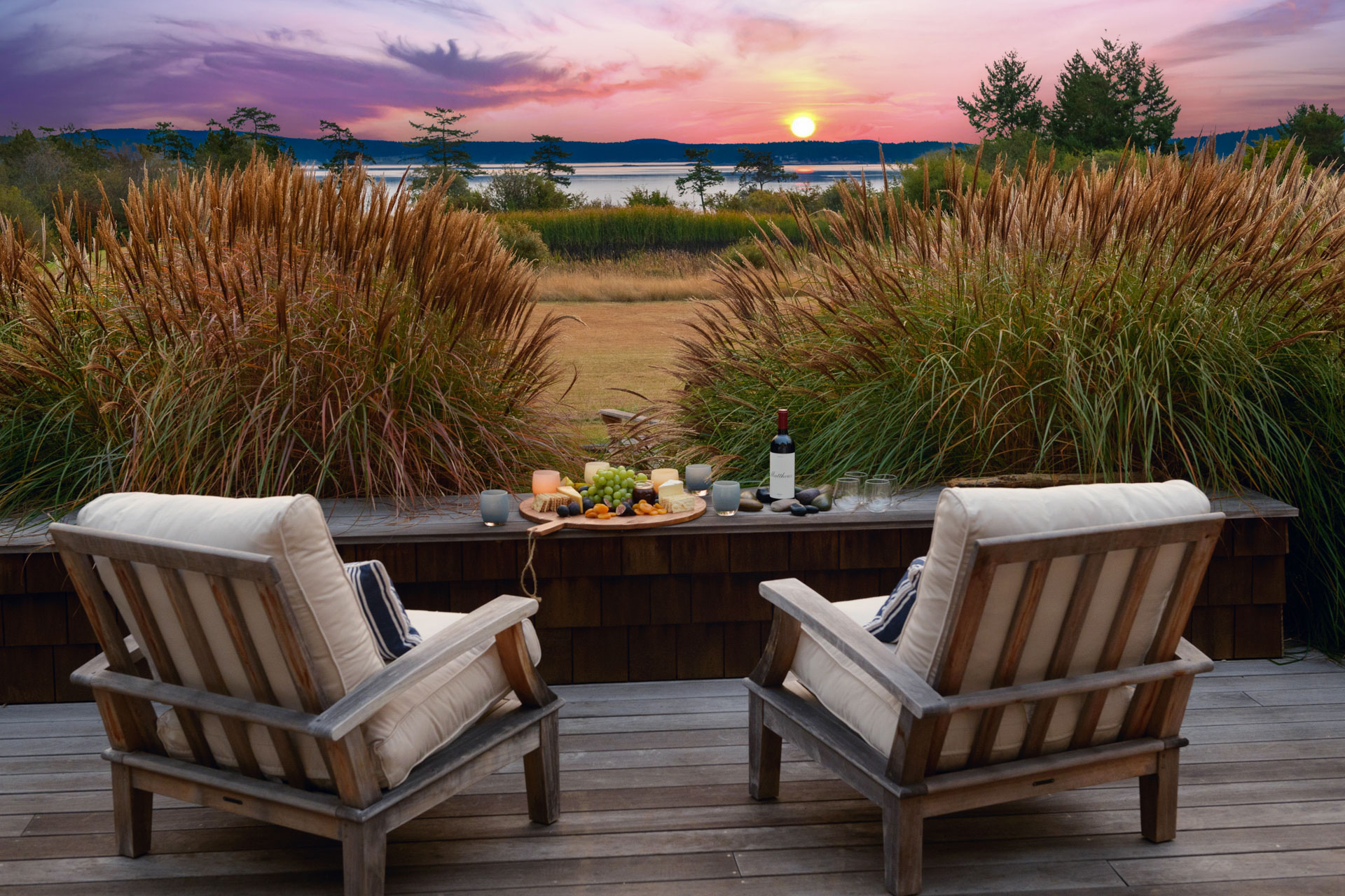 Two cushioned patio chairs sit on a wooden deck, facing a stunning sunset over a serene lake. Between the chairs is a table with a bottle of wine, glasses, and a platter of snacks. Tall grasses border the deck, enhancing the peaceful ambiance.