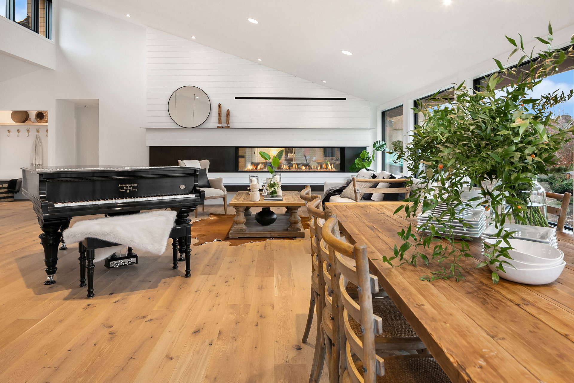 A modern open-concept living area featuring a grand piano, contemporary furniture, a long wooden dining table with chairs, and a green plant centerpiece. The space boasts a minimalist design with large windows, light wooden floors, and a sleek fireplace.