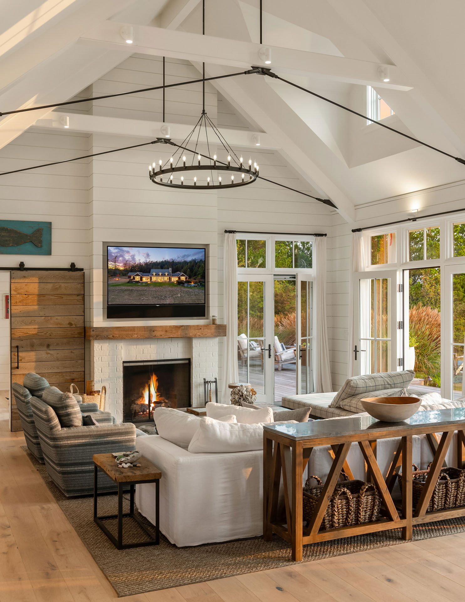 A bright, open living room with exposed beam ceilings, large windows, and a cozy fireplace. The space features a white sectional sofa, a wooden coffee table, and a flat-screen TV mounted above the fireplace. Natural light enhances the airy atmosphere.